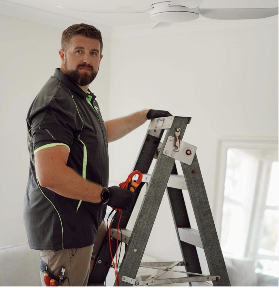 A man standing on a ladder working on a ceiling light fixture in a room with white walls and windows.