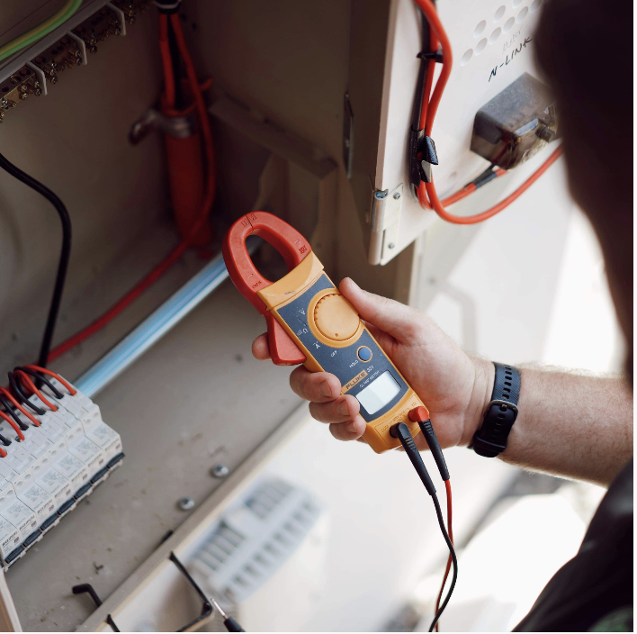 A person using a digital multimeter to measure electrical voltage inside an electrical panel with wires and circuit components.