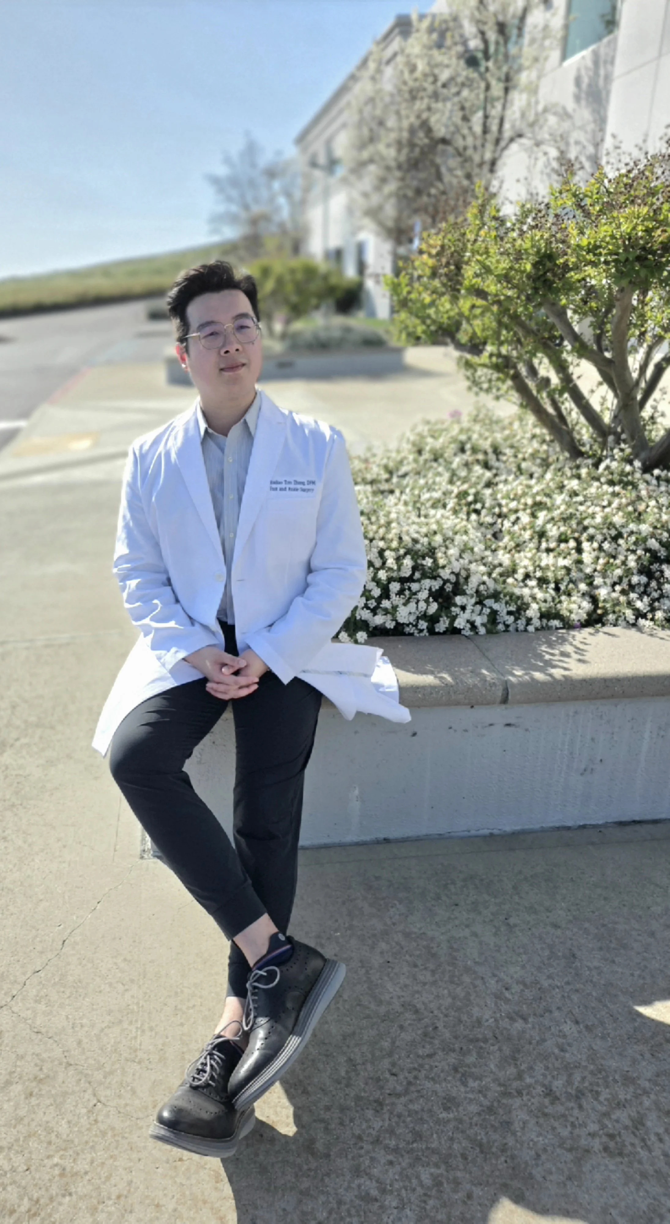 A young man wearing glasses and a white medical coat sitting on a concrete ledge with white flowers and greenery in the background.