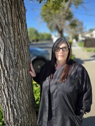 Woman with glasses and dark hair leaning against a tree on a sunny day in a residential neighborhood.