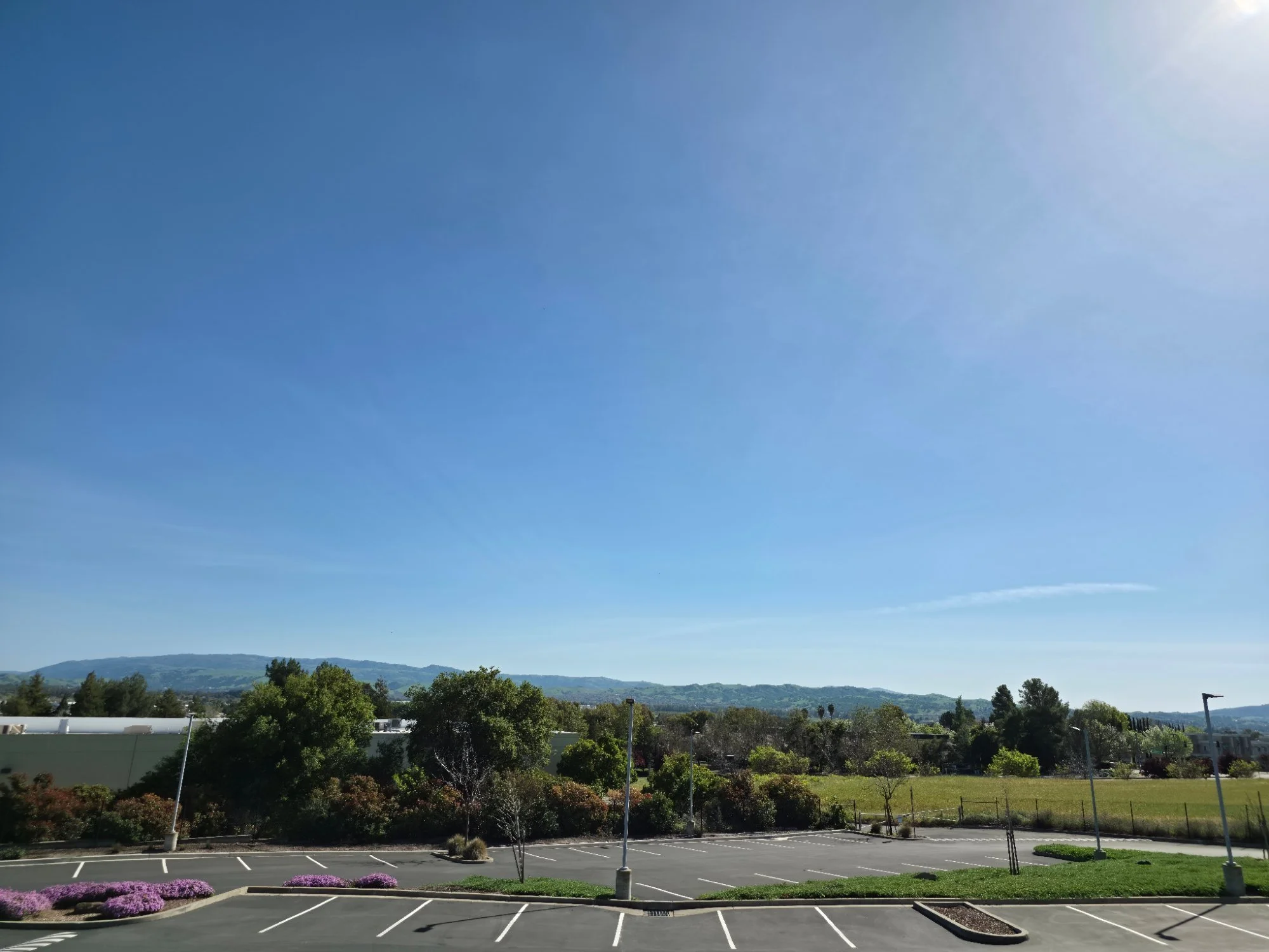 Empty outdoor parking lot with a backdrop of trees and distant hills under a clear blue sky.