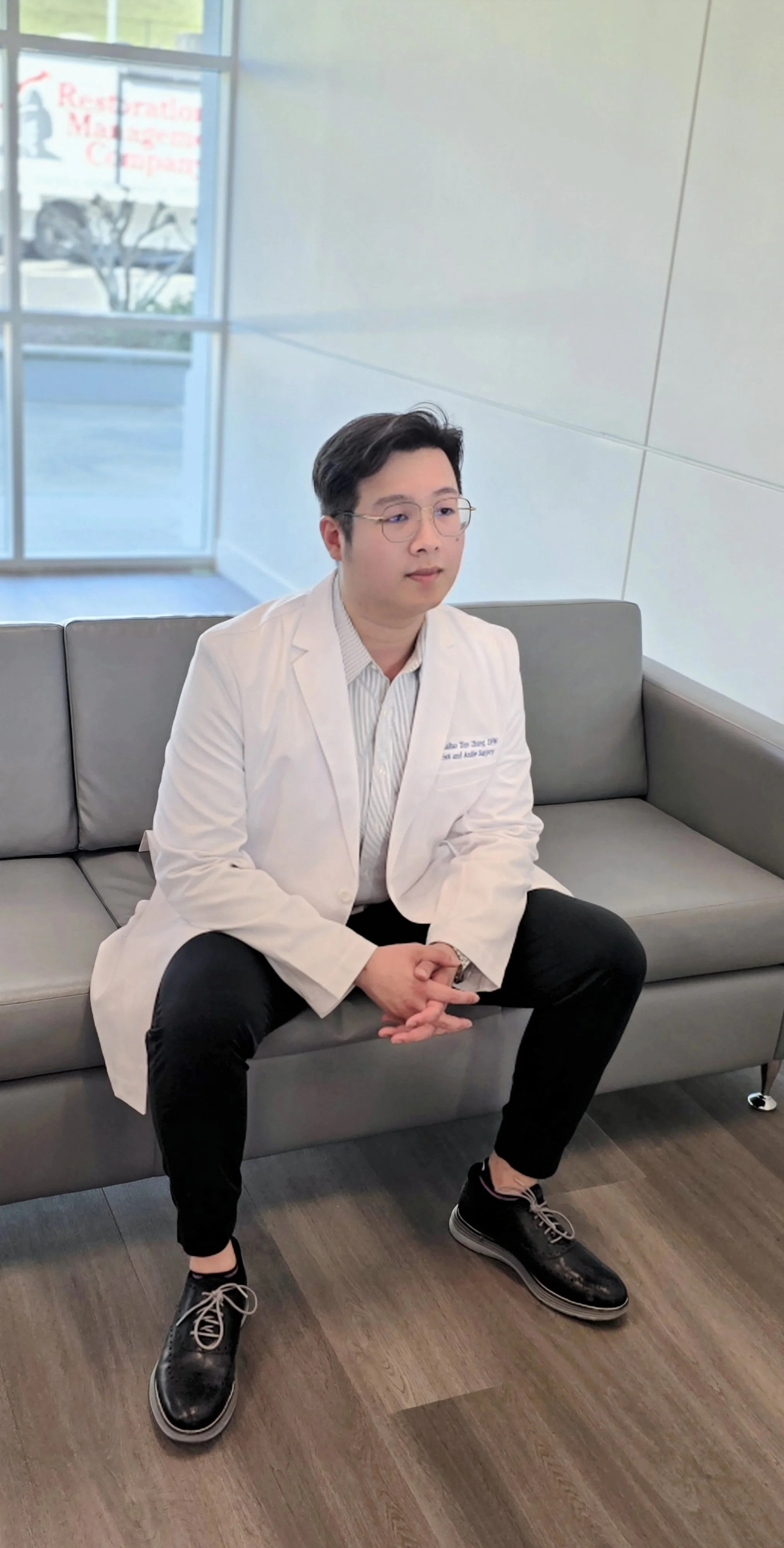 A young man with glasses, dressed in a white medical coat and dark pants, sitting on a gray sofa in a modern waiting area with wood flooring and large windows.