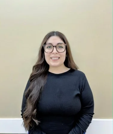 Woman with long dark hair and glasses smiling in front of a beige wall.