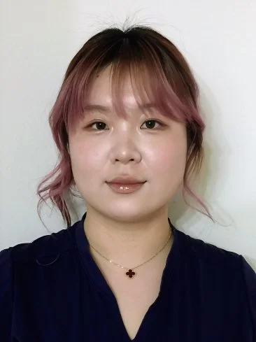 Young woman with short, wavy hair that is pink at the ends, wearing a navy top, a small flower-shaped necklace, and shiny lip gloss, standing against a plain white background.