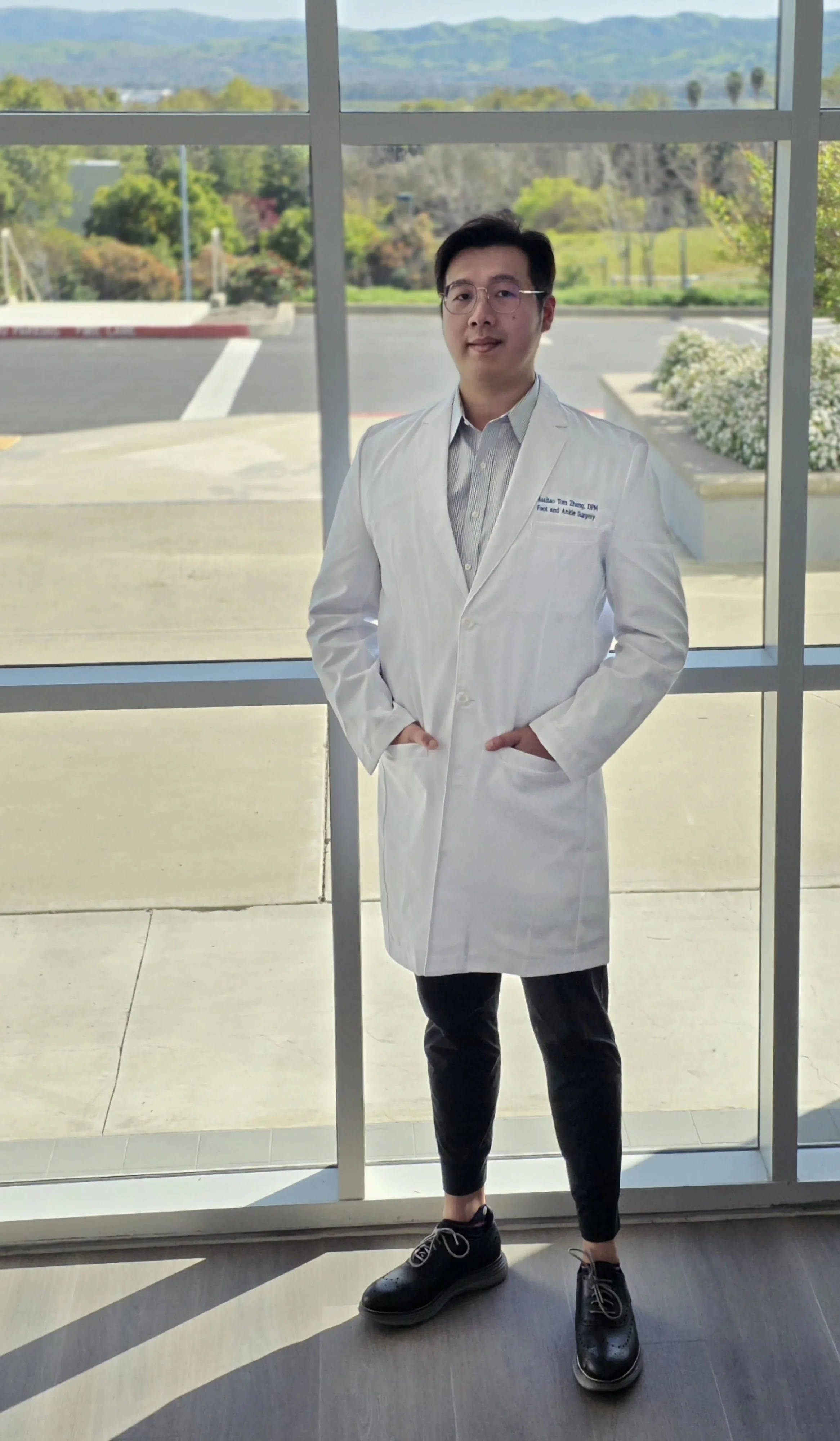 A man in a white doctor's coat standing inside in front of a large window, overlooking an outdoor scene with trees, mountains, and a parking lot.