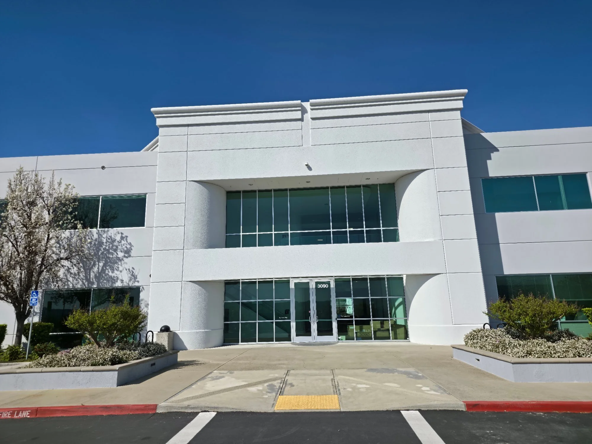 White commercial building with glass doors and windows, landscaped with small trees and flowers in front, under a clear blue sky.