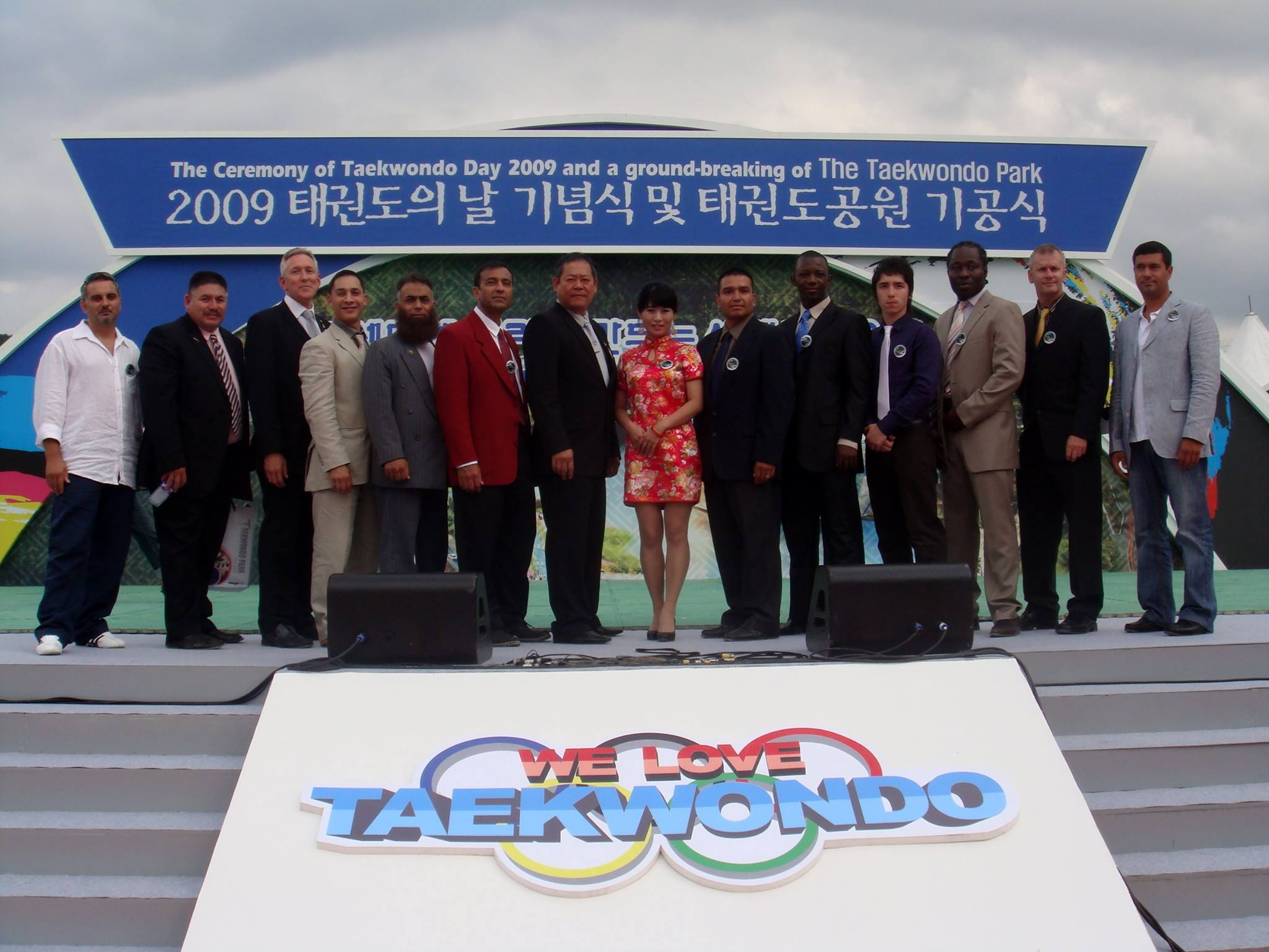 Group of people standing on a stage at an event celebrating Taekwondo, with a large sign in the background and a colorful 'We Love Taekwondo' banner in the foreground.