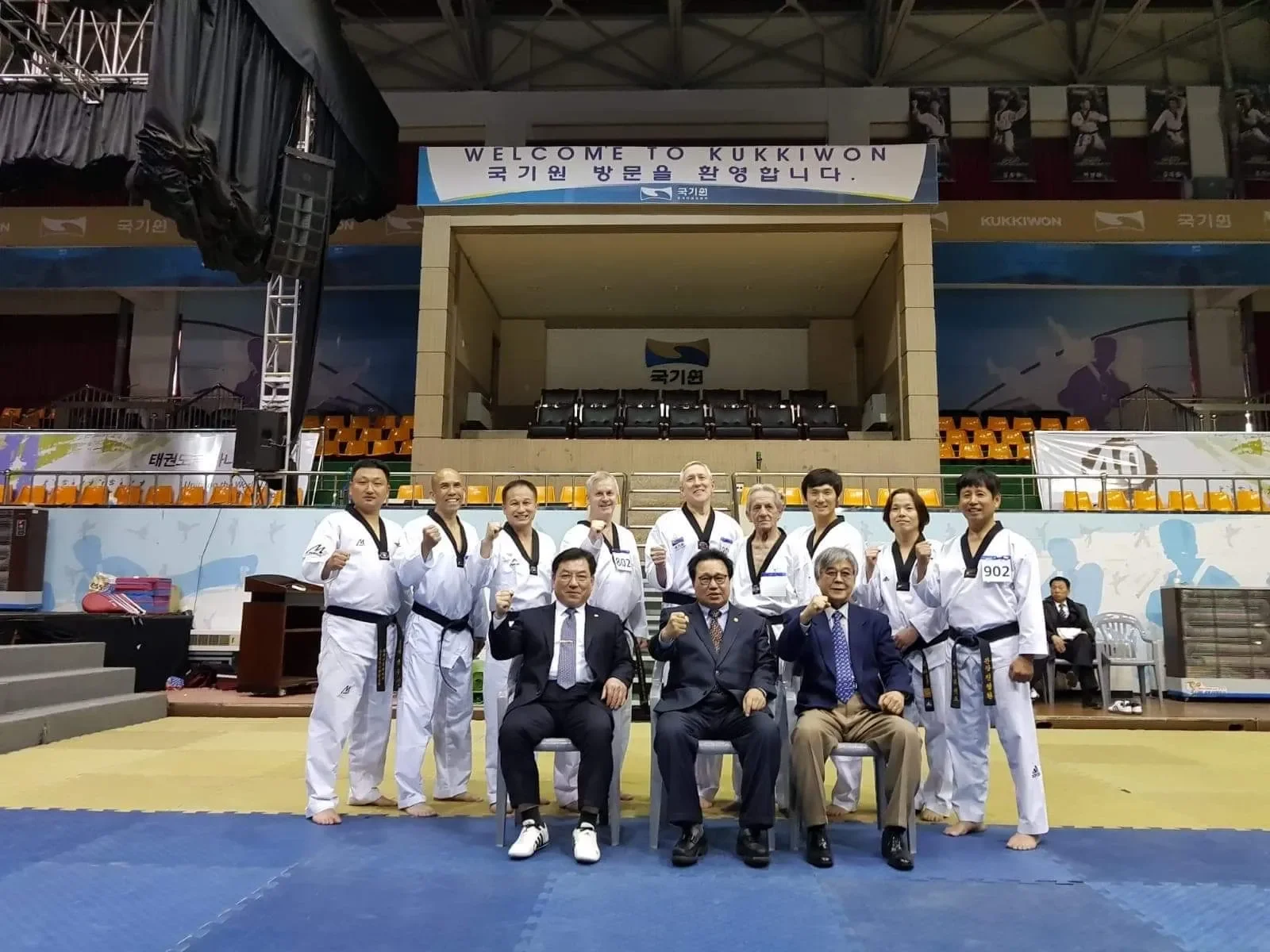 Group of taekwondo practitioners and officials posing for a photo in an indoor sports arena, with people in martial arts uniforms standing and seated in front of a sign that reads 'Welcome to Kukkiwon.'