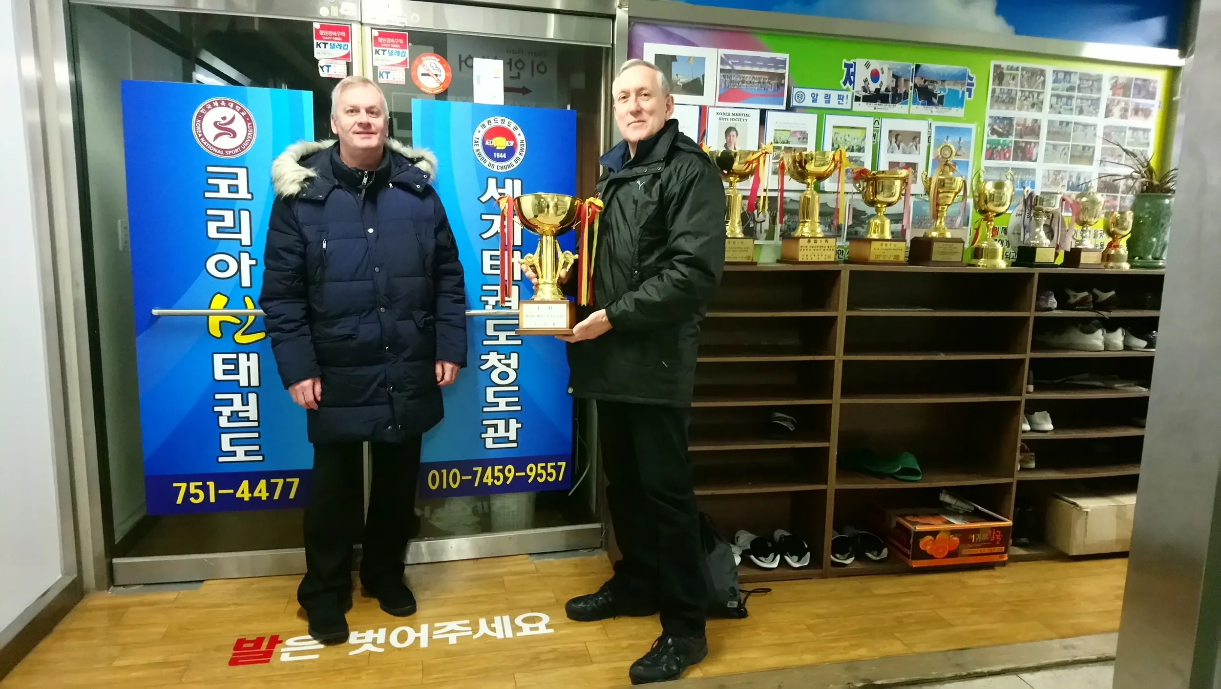 Two men stand inside an indoor sports facility, with one holding a trophy. Behind them are trophies on a shelf and colorful posters and photos on the wall. They are dressed in winter jackets, and the man on the right is wearing black sneakers.