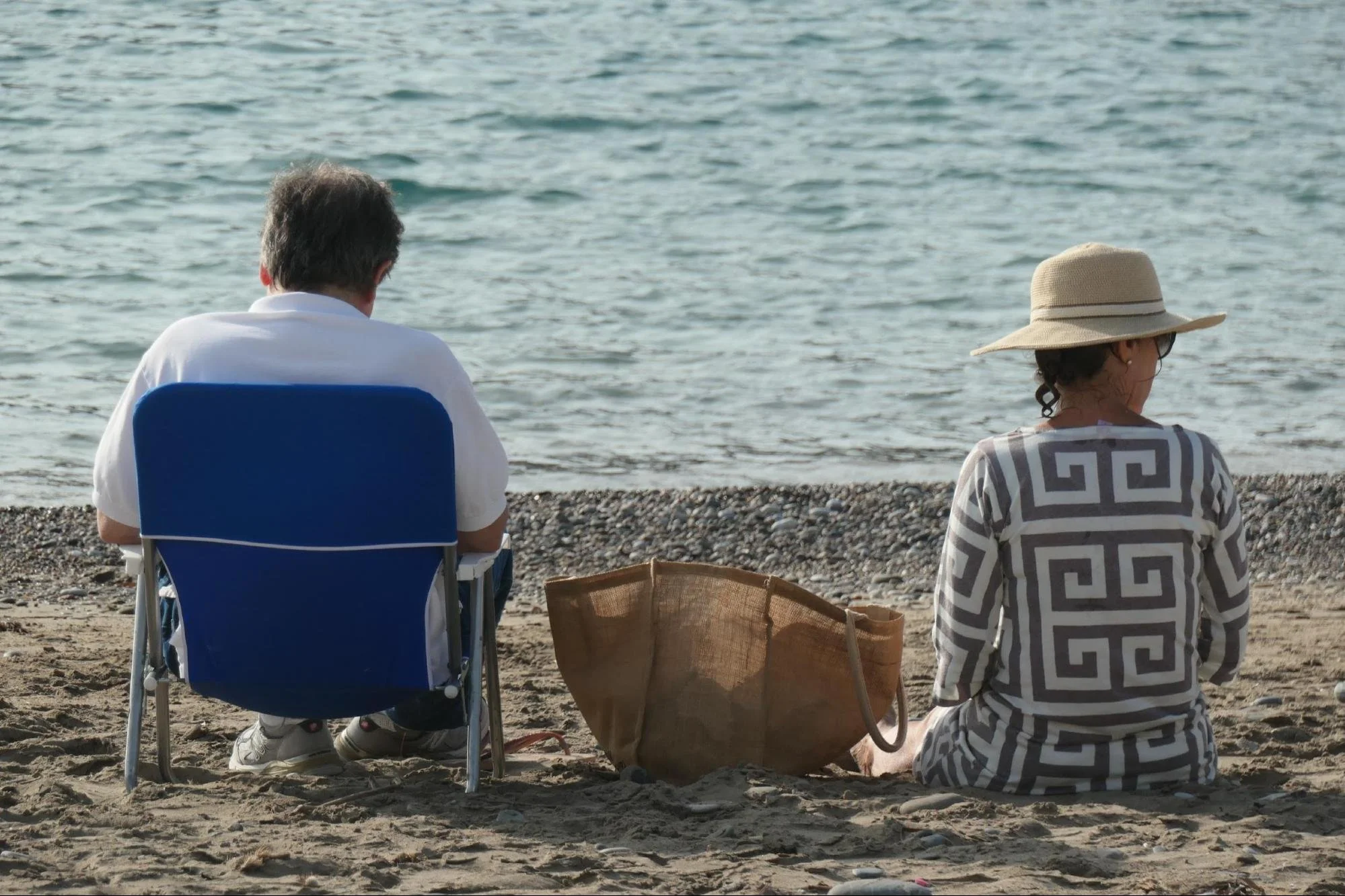 To personer sidder på stranden ved vandet, en mand på en blå stol og en kvinde med stor solhat, samt en stor taske ved siden af dem.