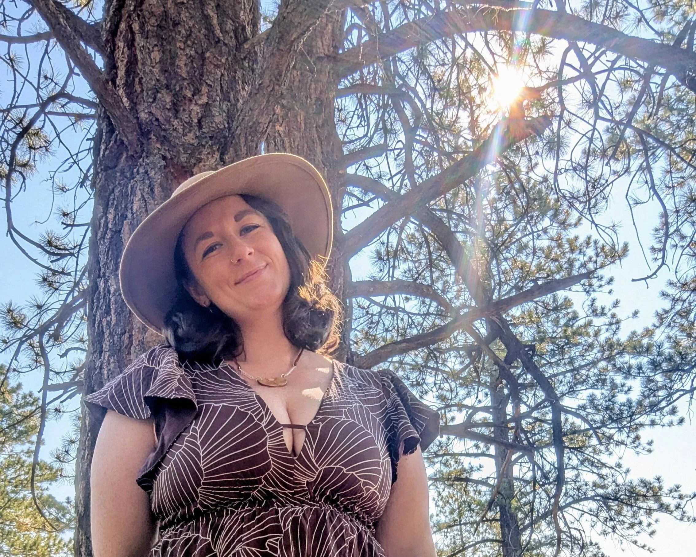 A woman with dark hair, wearing a beige wide-brimmed hat and a patterned black and white shirt, is standing outdoors in front of a large pine tree with the sun shining through its branches.