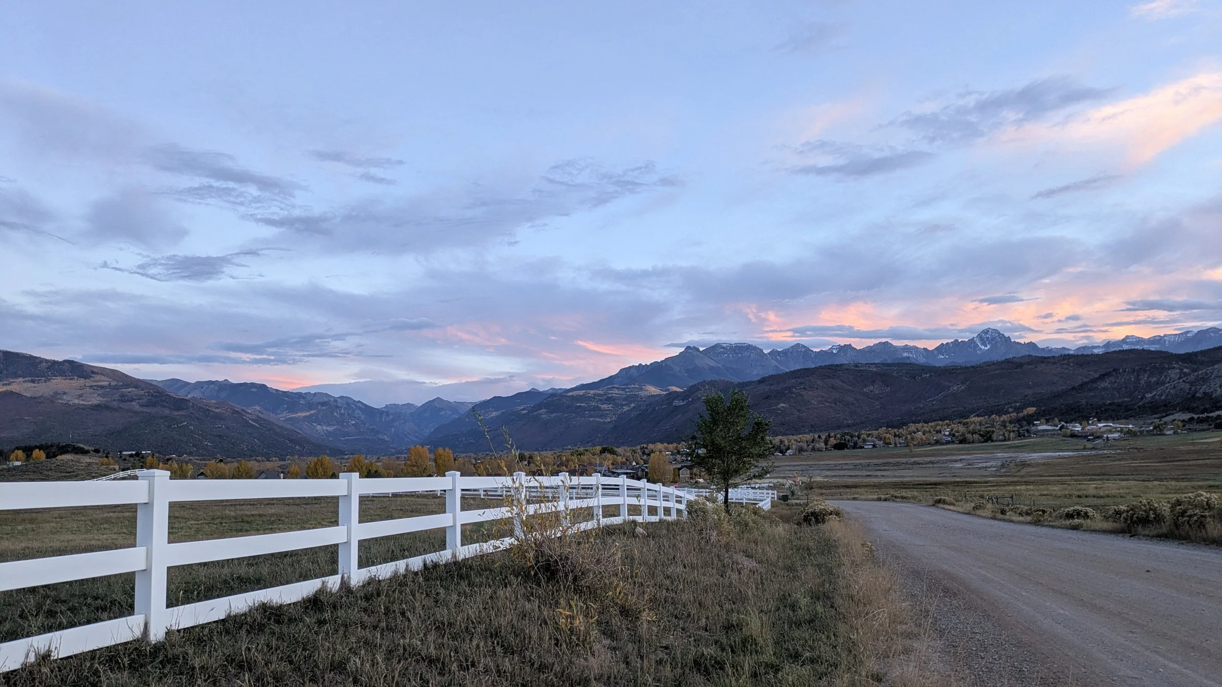 Scenic view of distant mountains under a cloudy sky during sunset, with a white fence in the foreground, a dirt road, and autumn trees in a rural landscape.