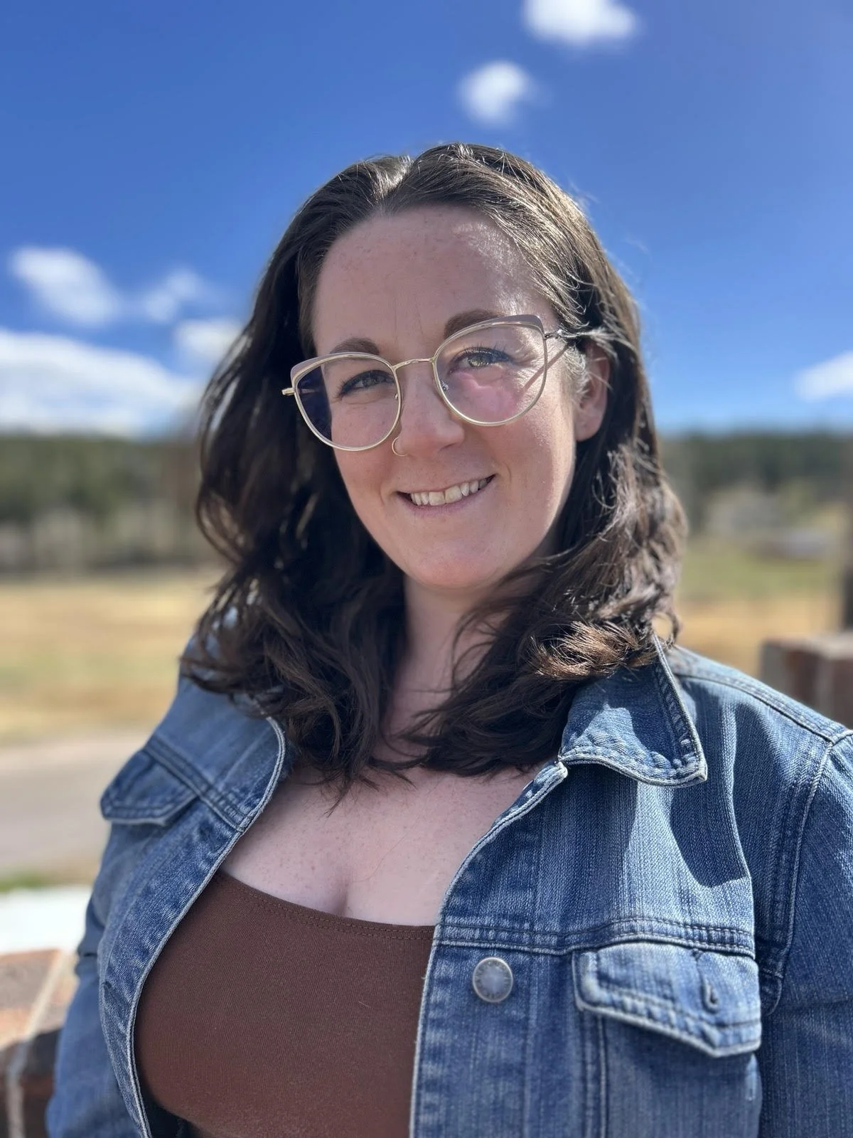 A woman smiling outdoors on a sunny day, wearing glasses and a denim jacket.