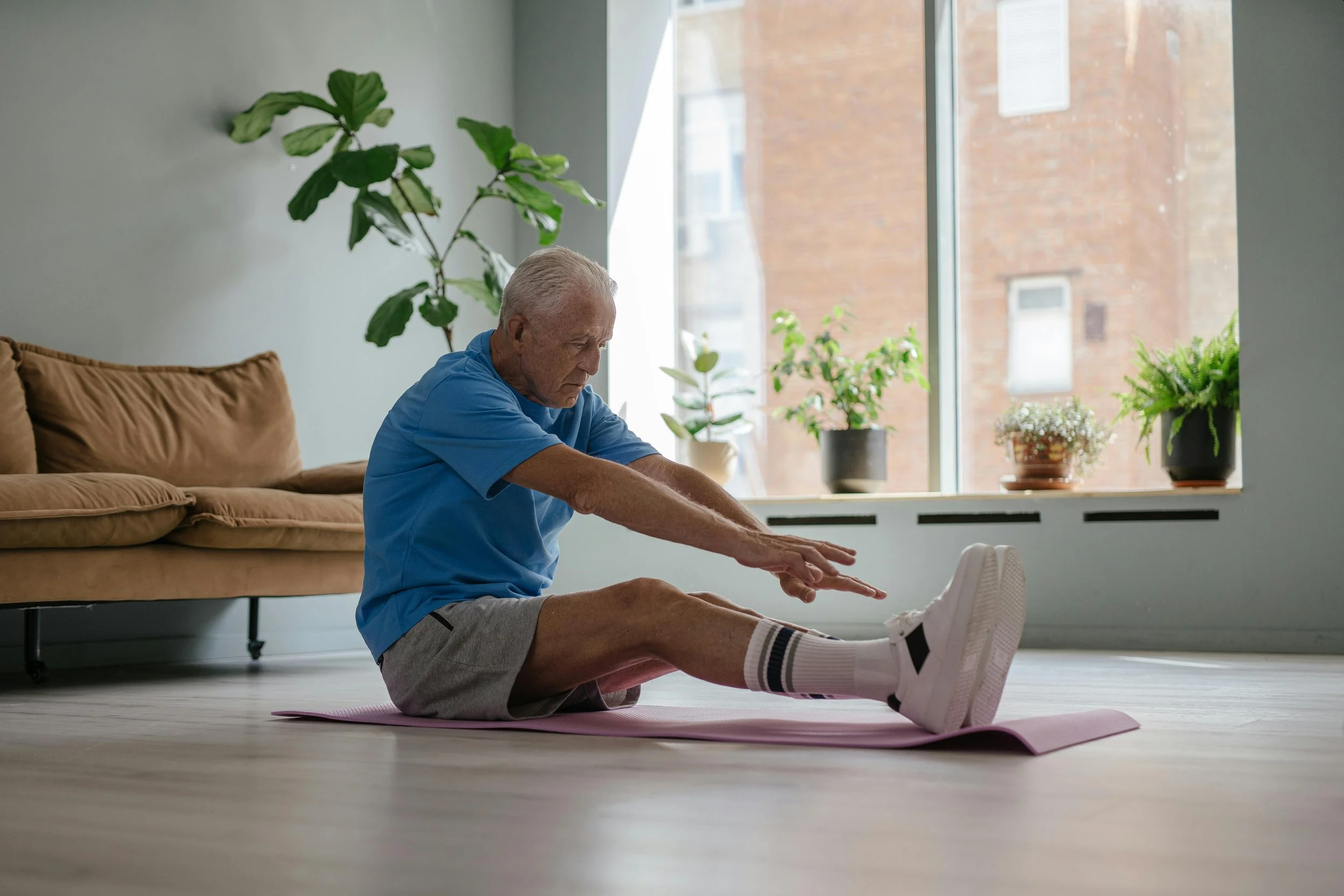 An elderly man doing stretching exercises on a pink yoga mat in a living room with large window and potted plants.