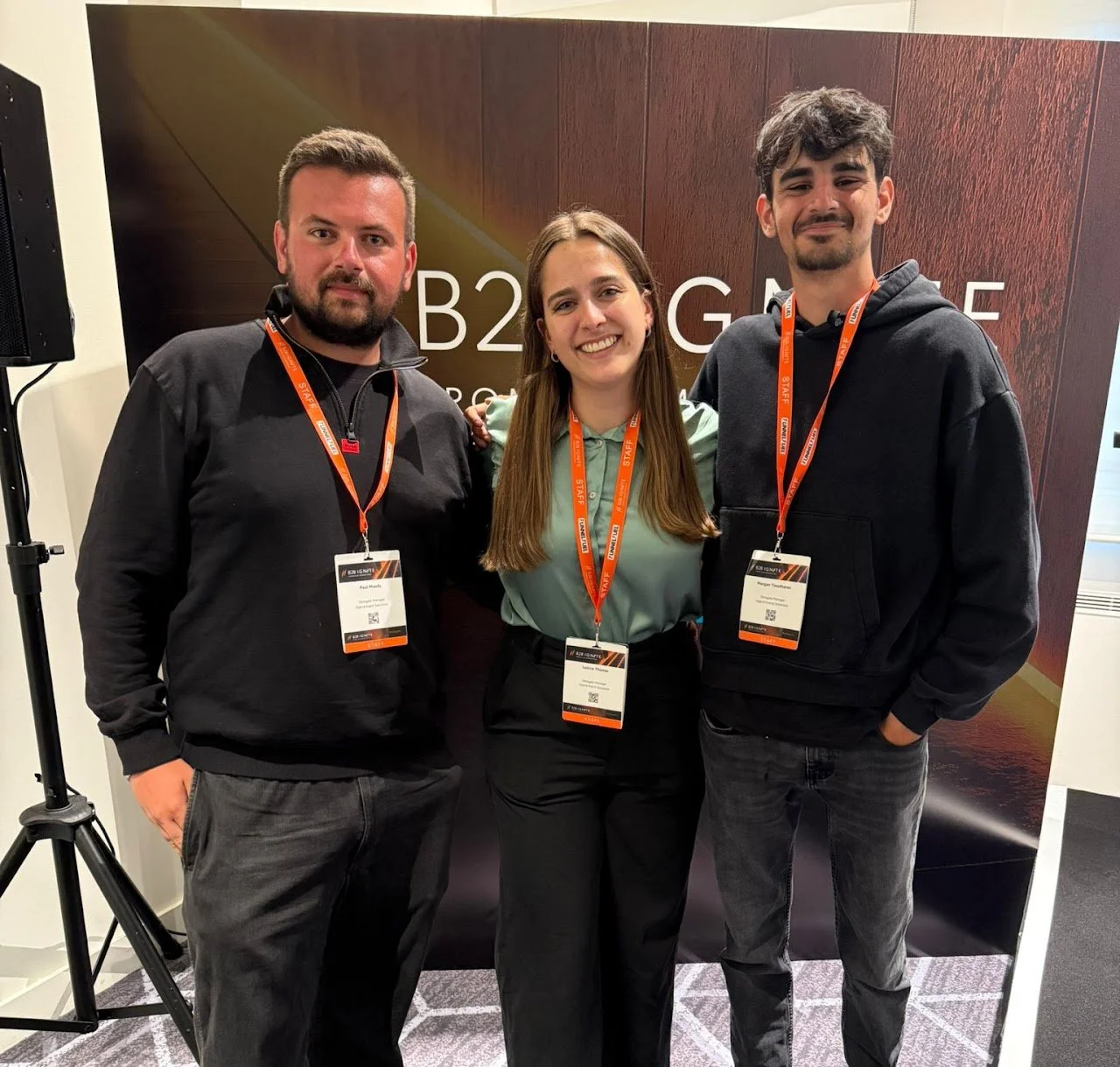 Event coordinator standing with colleagues at a corporate conference, wearing event badges and smiling in front of a branded exhibition booth.