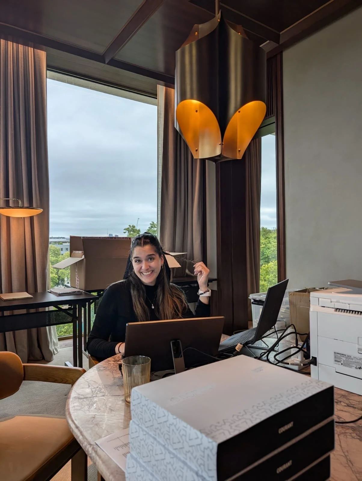 A woman with long dark hair smiling at the camera, sitting at a marble table with laptops, a glass of water, and a printer in a modern room with large window, curtains, and a unique gold and black chandelier.