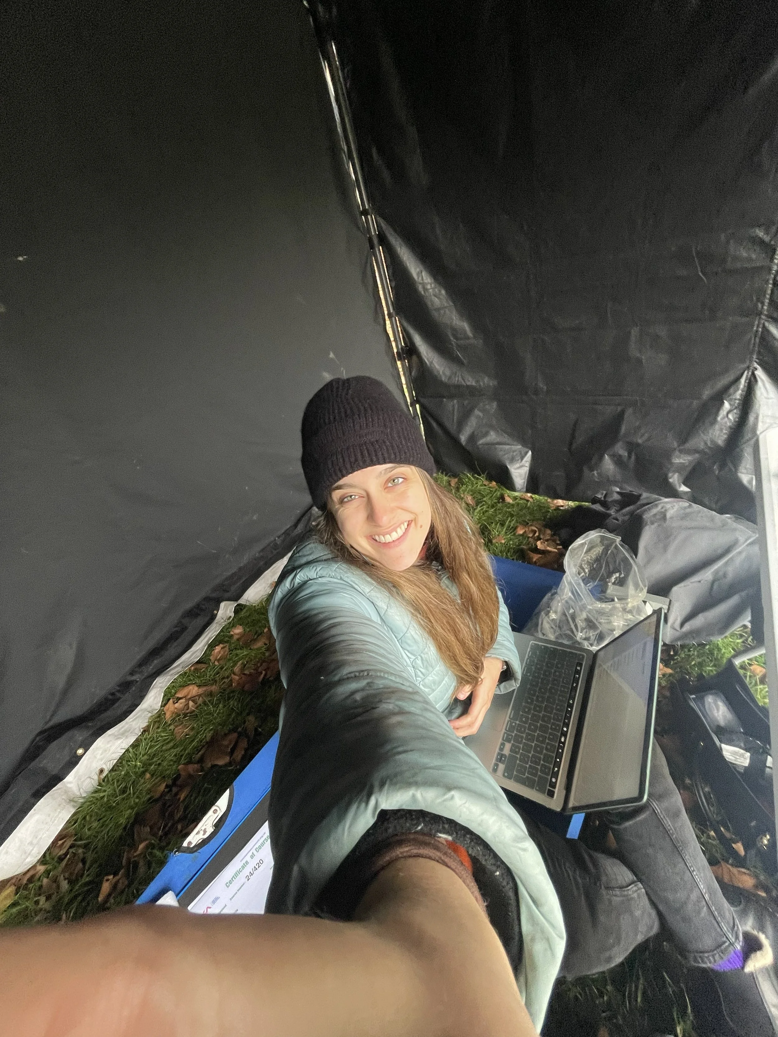 A woman taking a selfie from an elevated perspective inside a small outdoor tent or shelter. She is smiling, wearing a black beanie, a light gray jacket, and has long brown hair. She is sitting on the ground next to a laptop and some papers, with a black bag nearby, and the tent floor is covered with grass and fallen leaves.