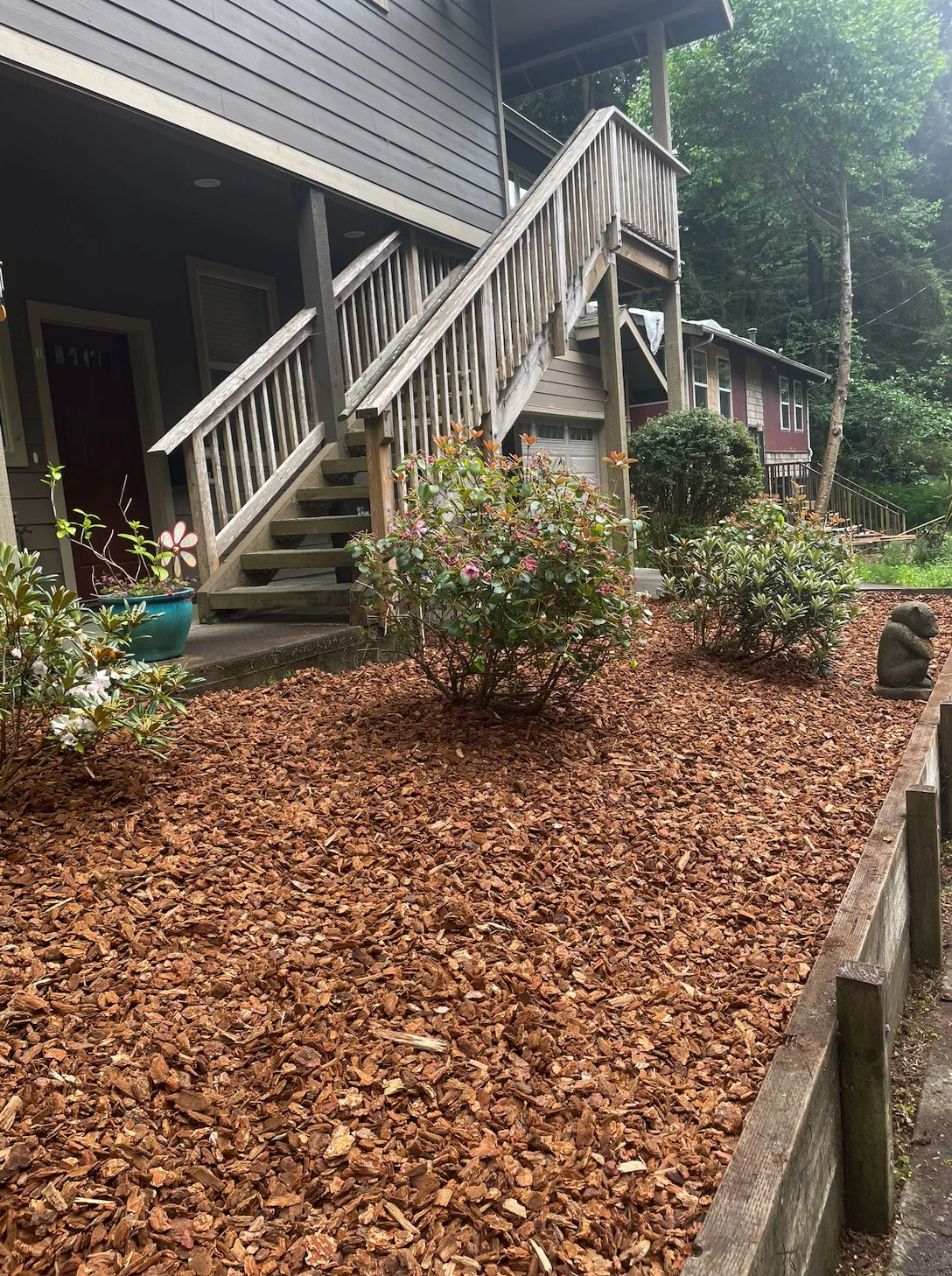 A residential backyard with a mulched ground, a staircase leading to a house door, several potted plants, shrubs, trees, and a decorative stone sculpture.
