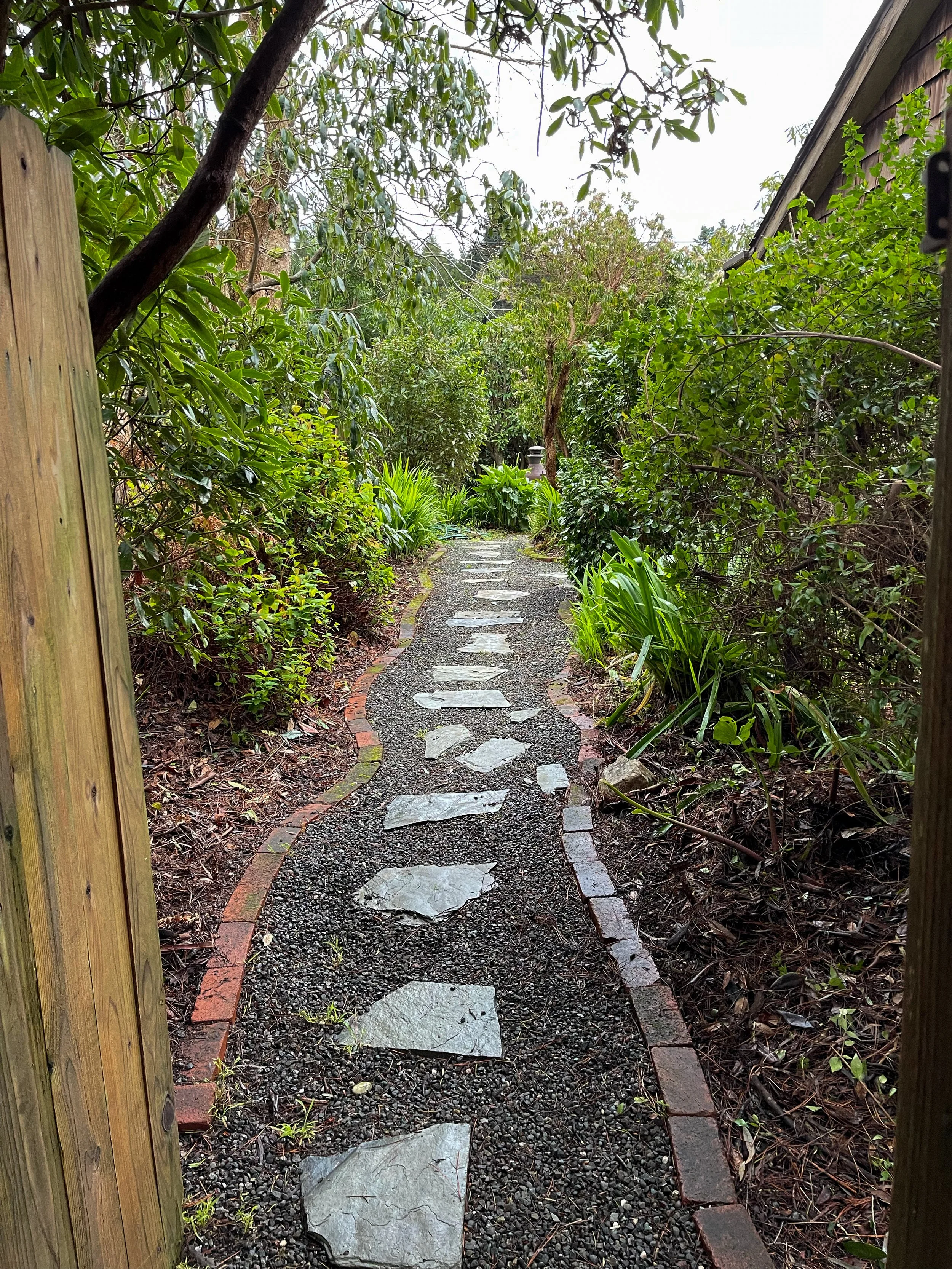 A narrow garden pathway made of irregular grey stones and bordered by red bricks, winding through lush green shrubbery and trees.