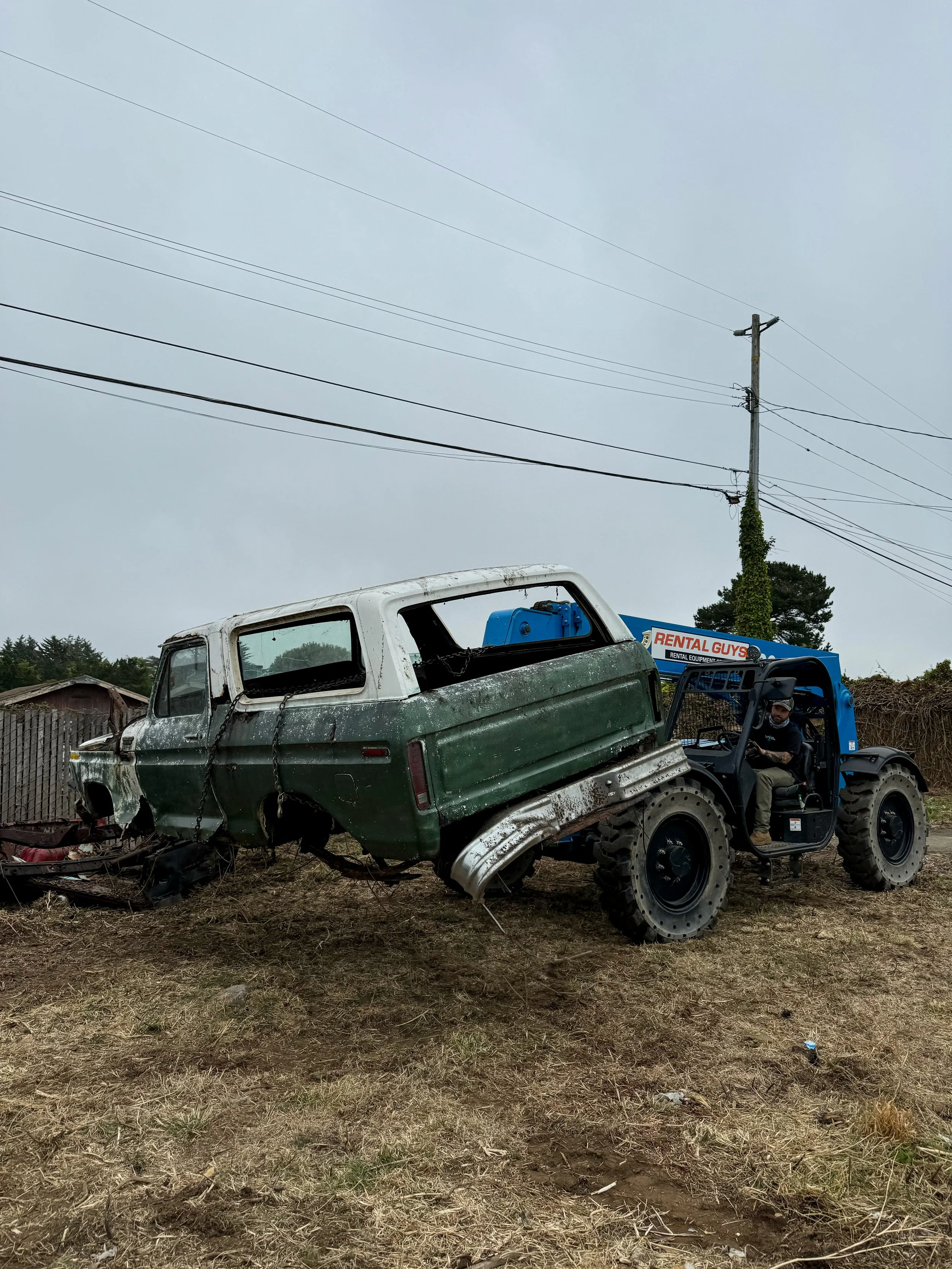 A blue tractor with a sign 'RENTAL GUYS' is lifting an old, damaged green and white vehicle off the ground. The vehicle appears to be an abandoned or wrecked station wagon with broken windows and rusted body, on a dirt field with overcast sky.