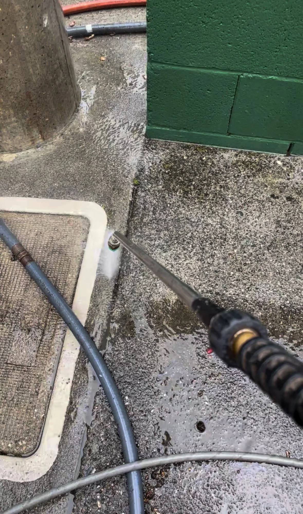 A pressure washer spraying water on a concrete sidewalk near a drain grate, with hoses and a green painted brick wall in the background.