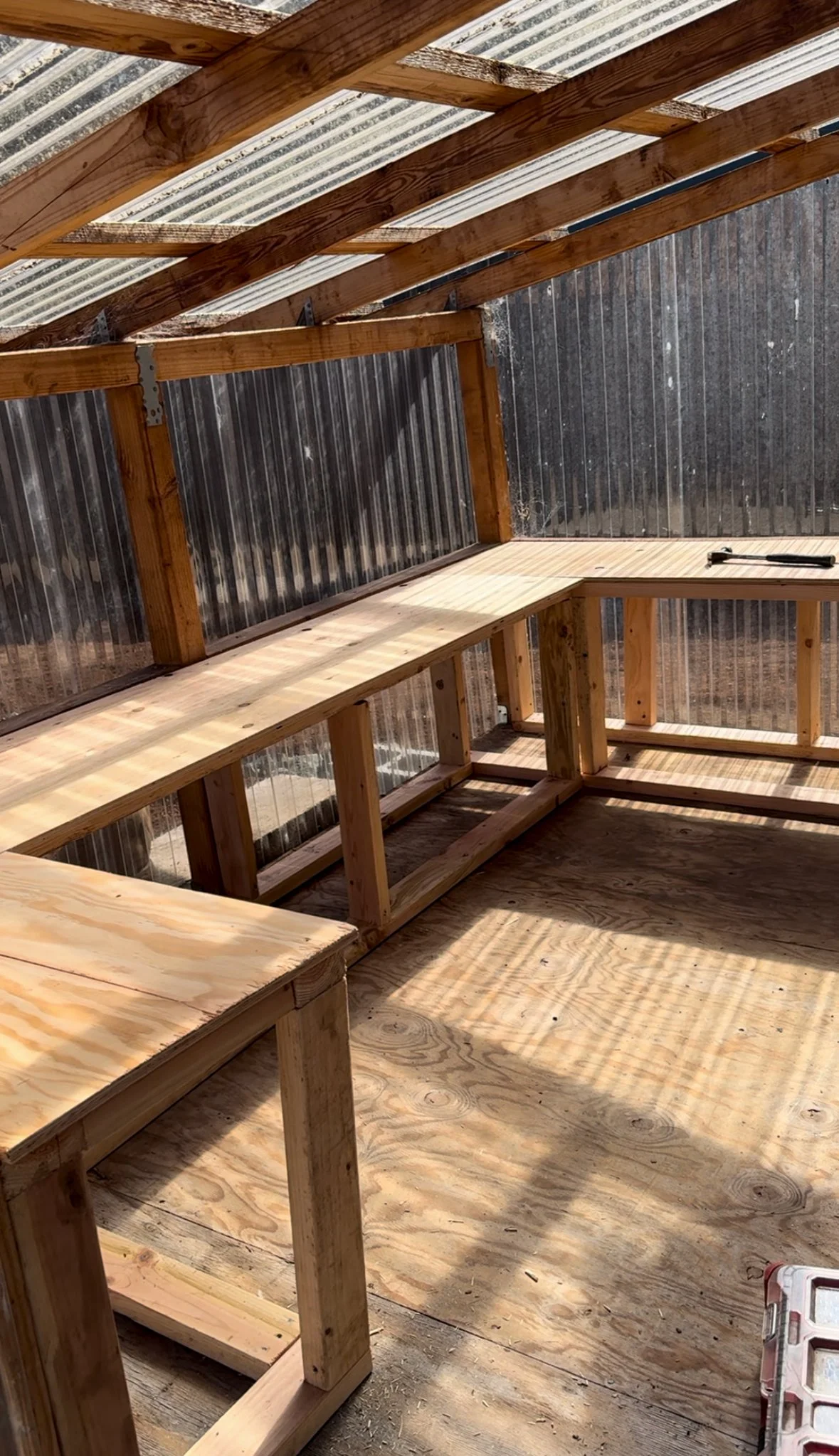 Unfinished wooden workbench inside a metal-walled shed with sunlight casting shadows on the plywood floor.