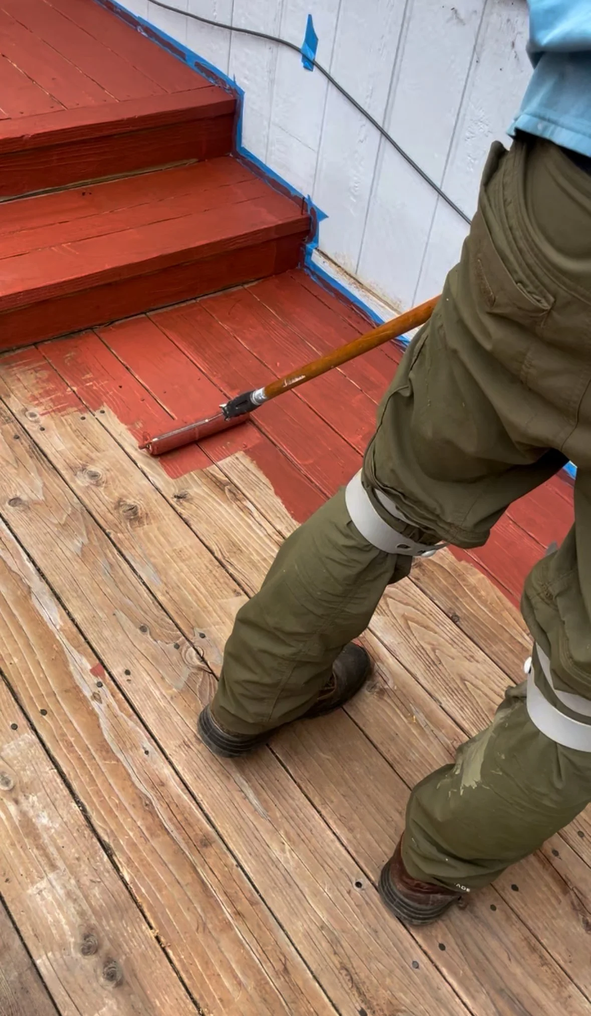 Person staining a wooden deck with a roller brush, wearing brown pants, a blue shirt, and brown boots, near white tiled walls and stairs.