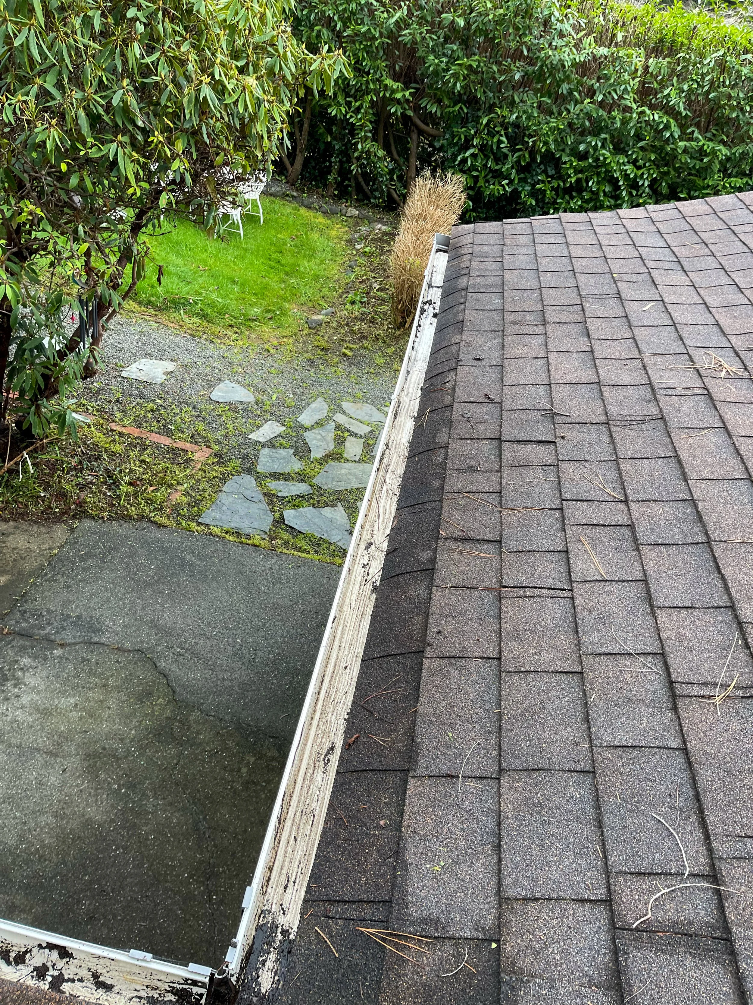 View of a rooftop with dark shingles, a white gutter, and a backyard with a stone pathway, green grass, bushes, and a small patch of grass with a white outdoor table and chairs.