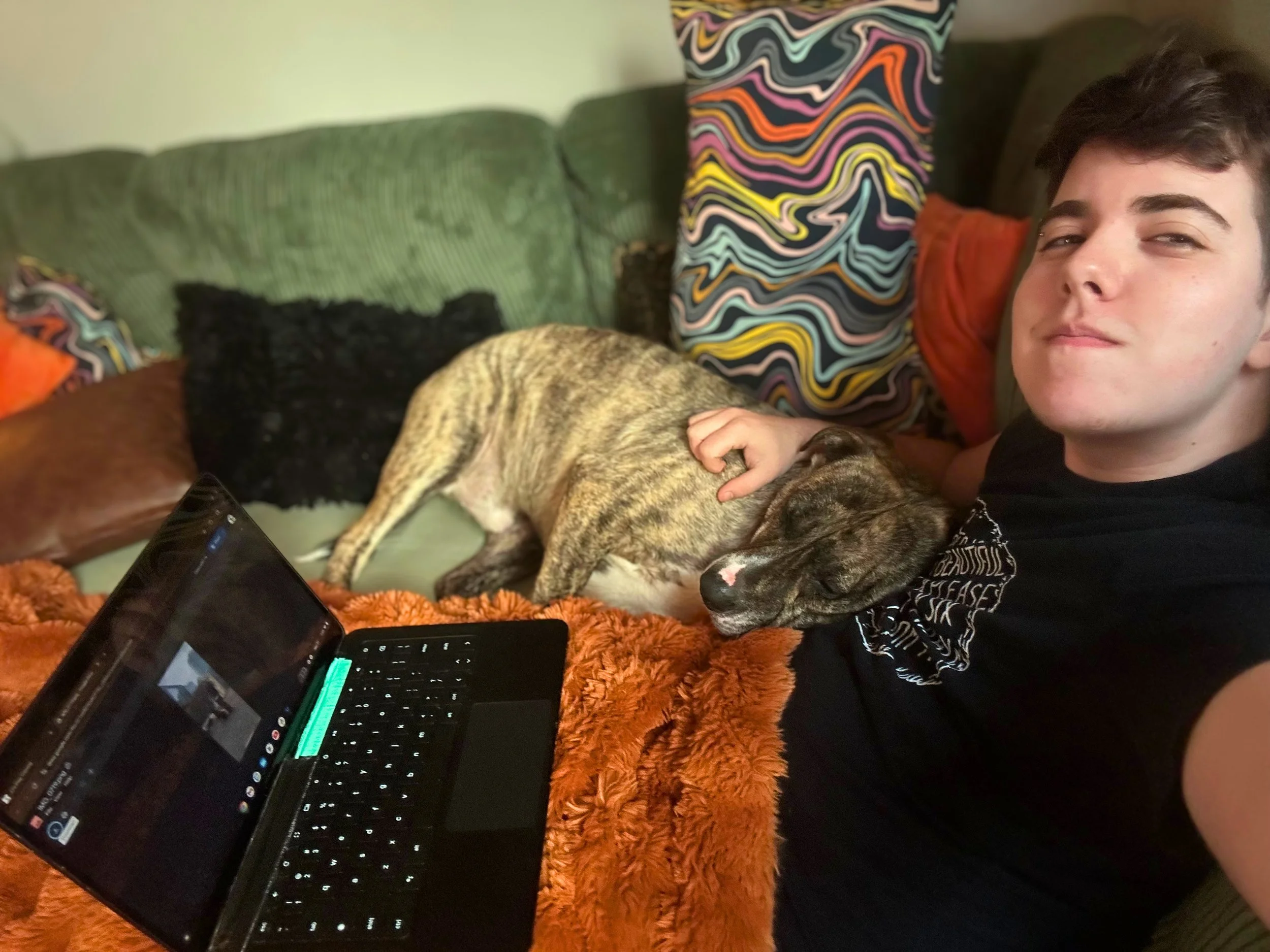 A young man with short brown hair is taking a selfie on a couch with a brindle-colored dog resting its head on his chest, surrounded by colorful pillows.