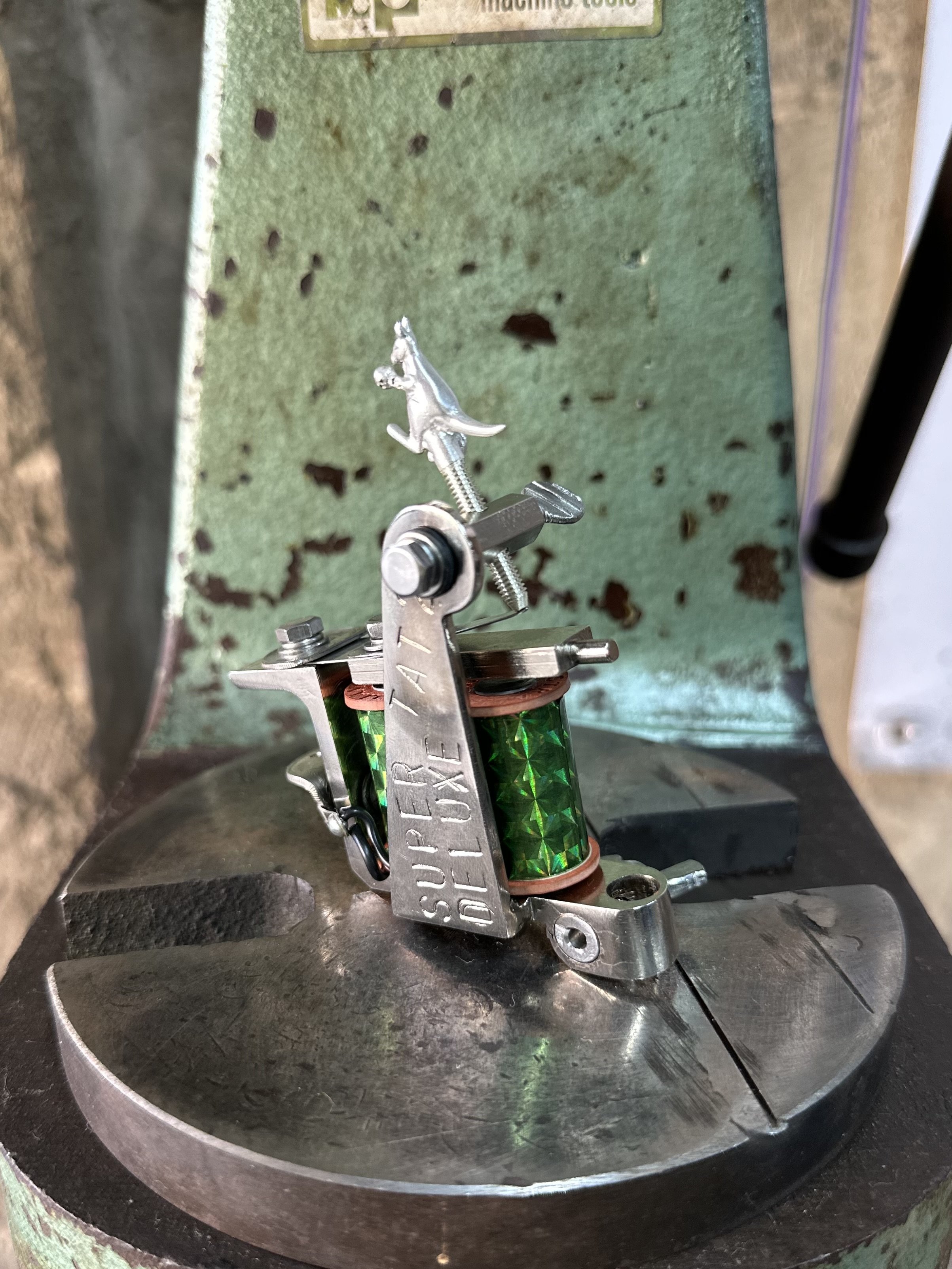 A close-up of a machine with a metal clamp and a green spool of wire, mounted in a circular metal base with a rusty surface. The background shows a green textured surface with small holes and rust spots.