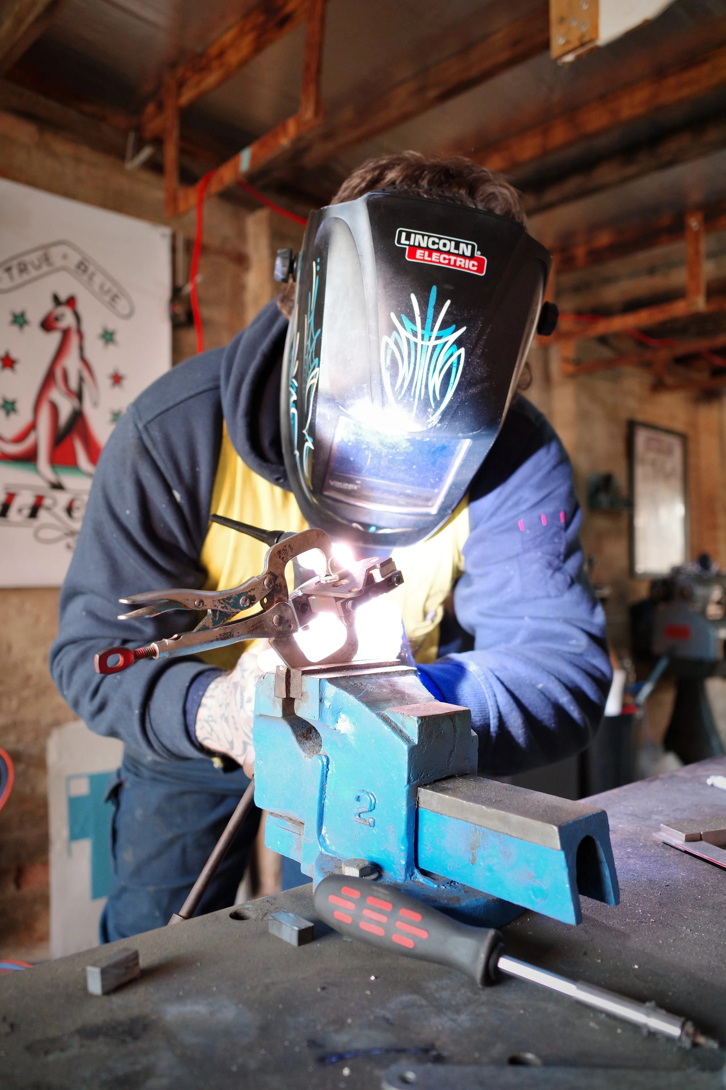 Person welding metal parts in a workshop, wearing a protective welding helmet with a design and the brand Lincoln Electric, using a welding tool on a metal piece clamped in a blue vise.