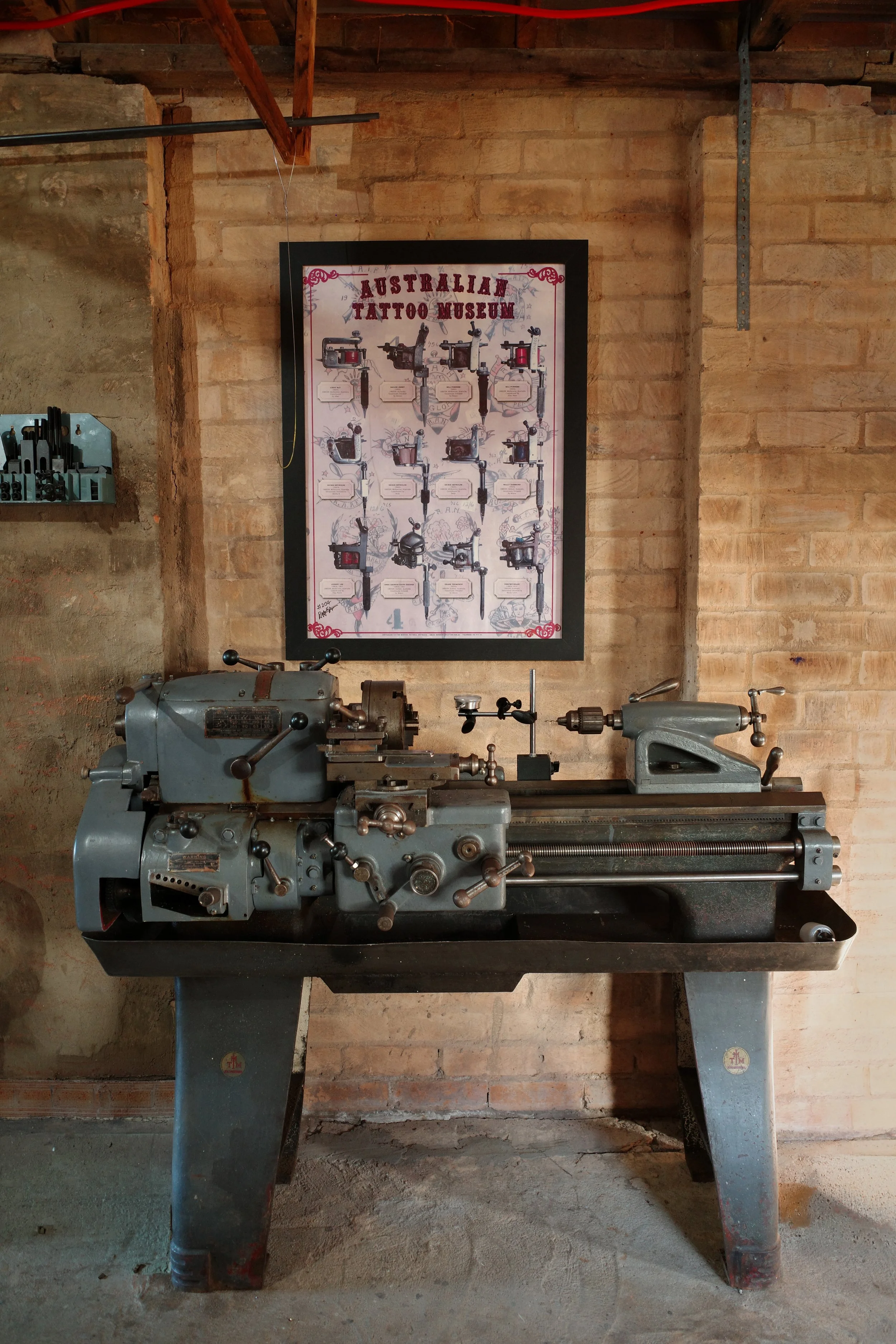 A vintage metal lathe machine in a workshop with a brick wall background, and a framed poster labeled 'Australian Tattoo Museum' hanging above it.