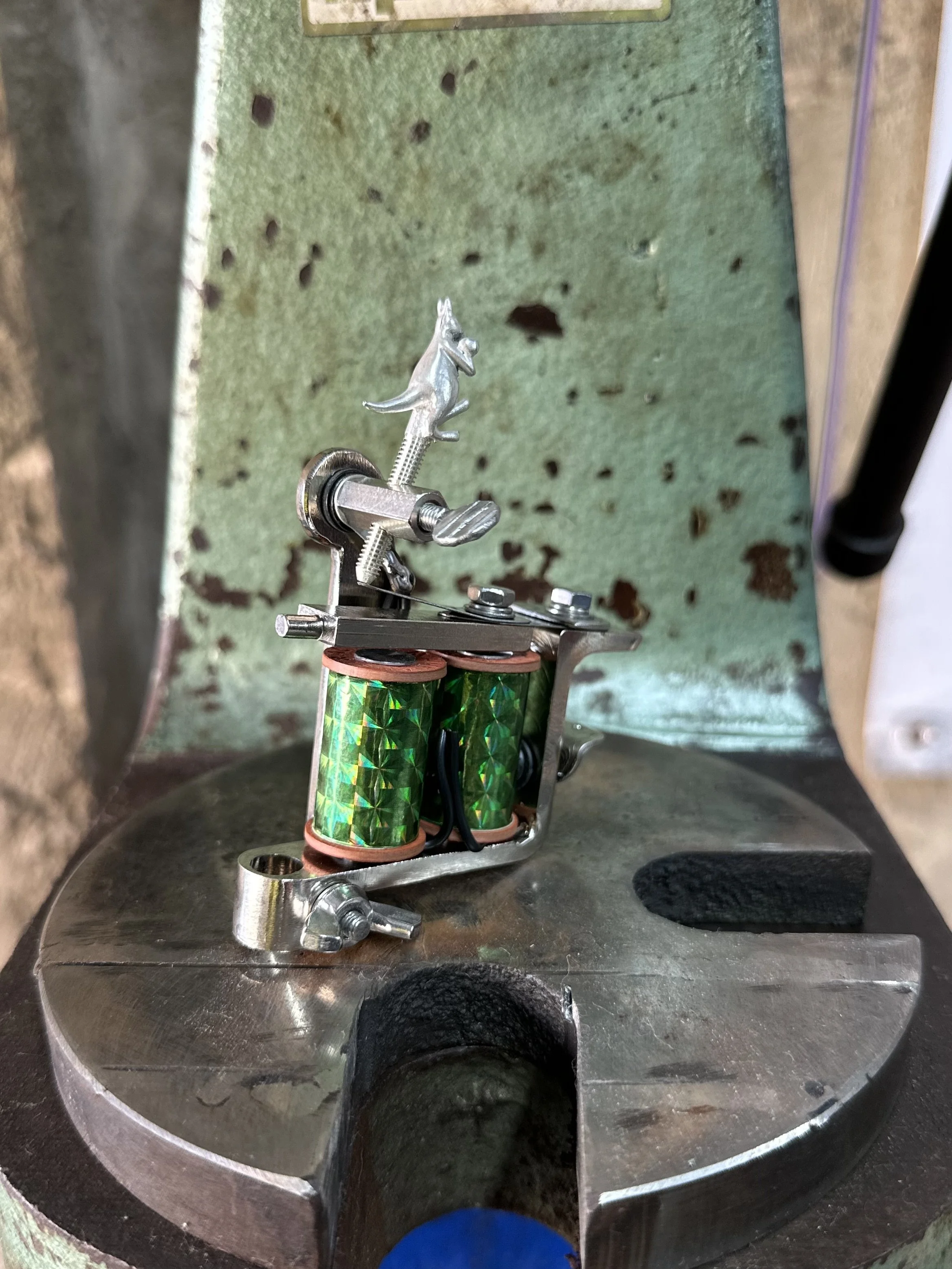Close-up of a mechanized machine with a small metal figure resembling a dinosaur on top of a spring, surrounded by components with shiny green and copper-colored coils, against a green and rusted background.