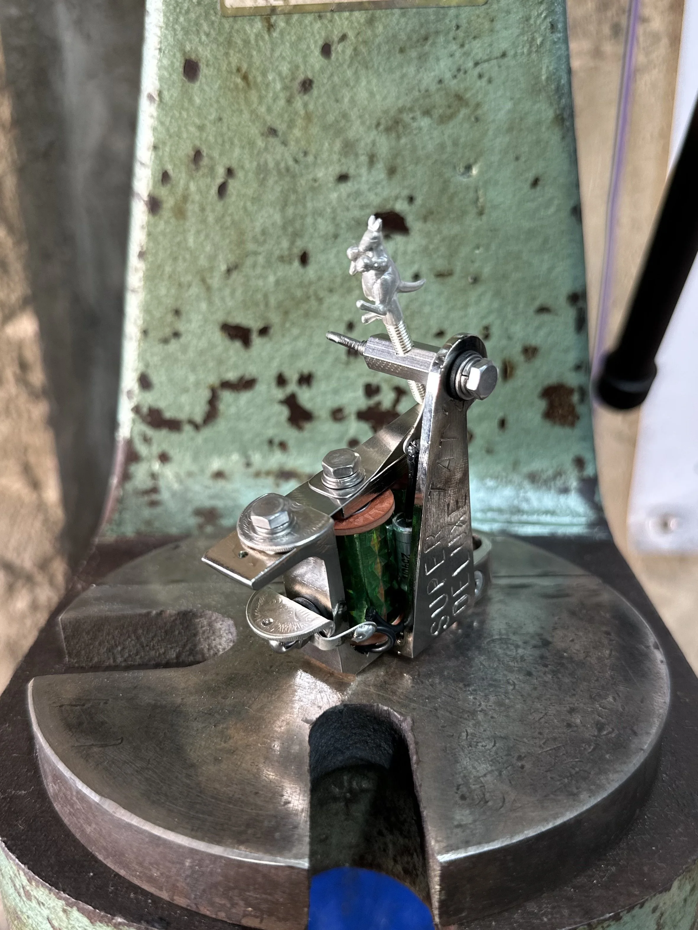 Close-up of a metal cricket figurine mounted on a mechanical device inside a circular metallic structure. The background is a worn, green wall with holes and spots.
