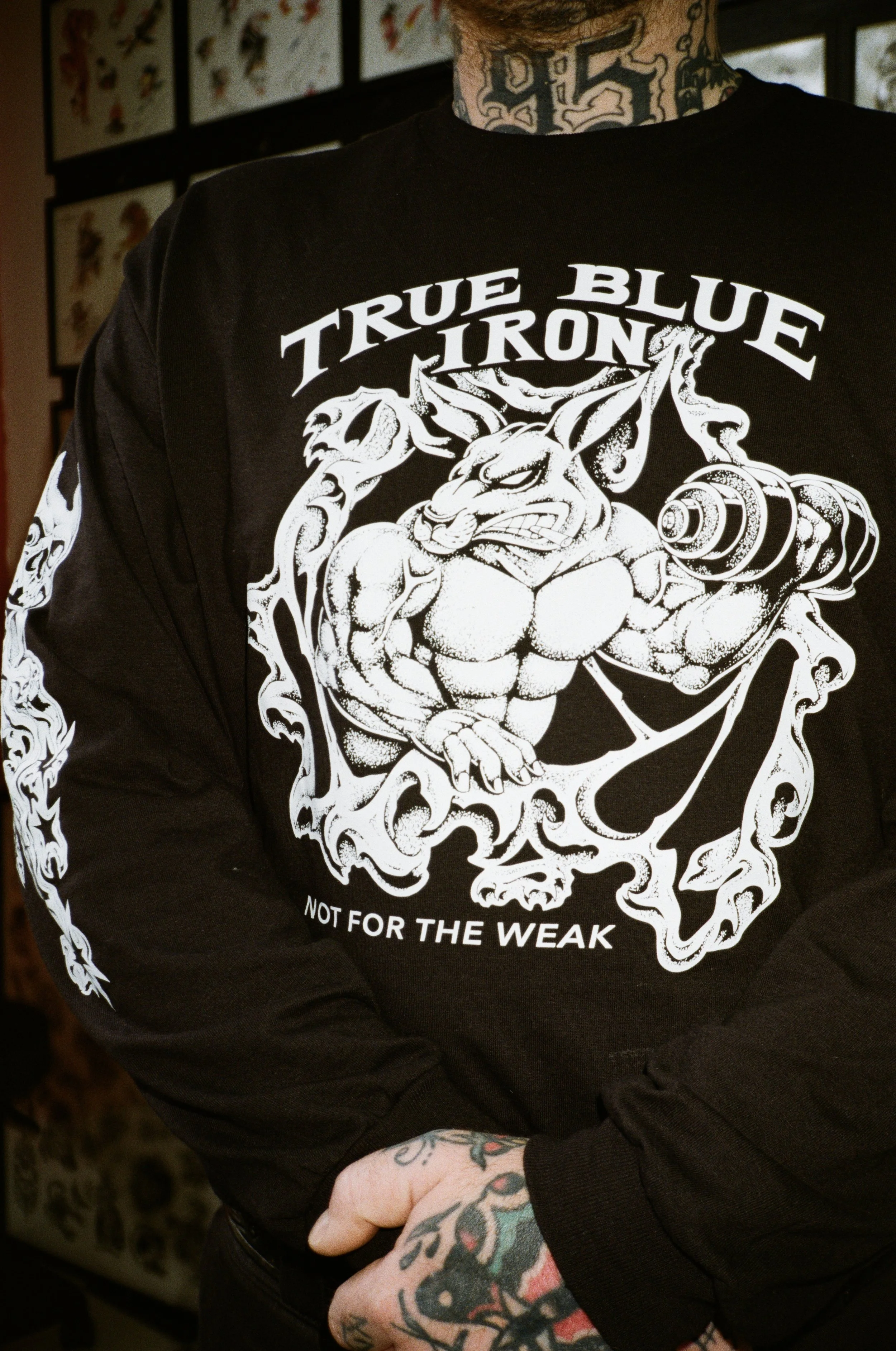 Person wearing a black long-sleeve shirt with a white graphic design of a muscular creature holding a barbell, with the text "TRUE BLUE TRON" and "NOT FOR THE WEAK". The person has tattoos on neck and hands, and is standing indoors with framed pictures of tattoos in the background.