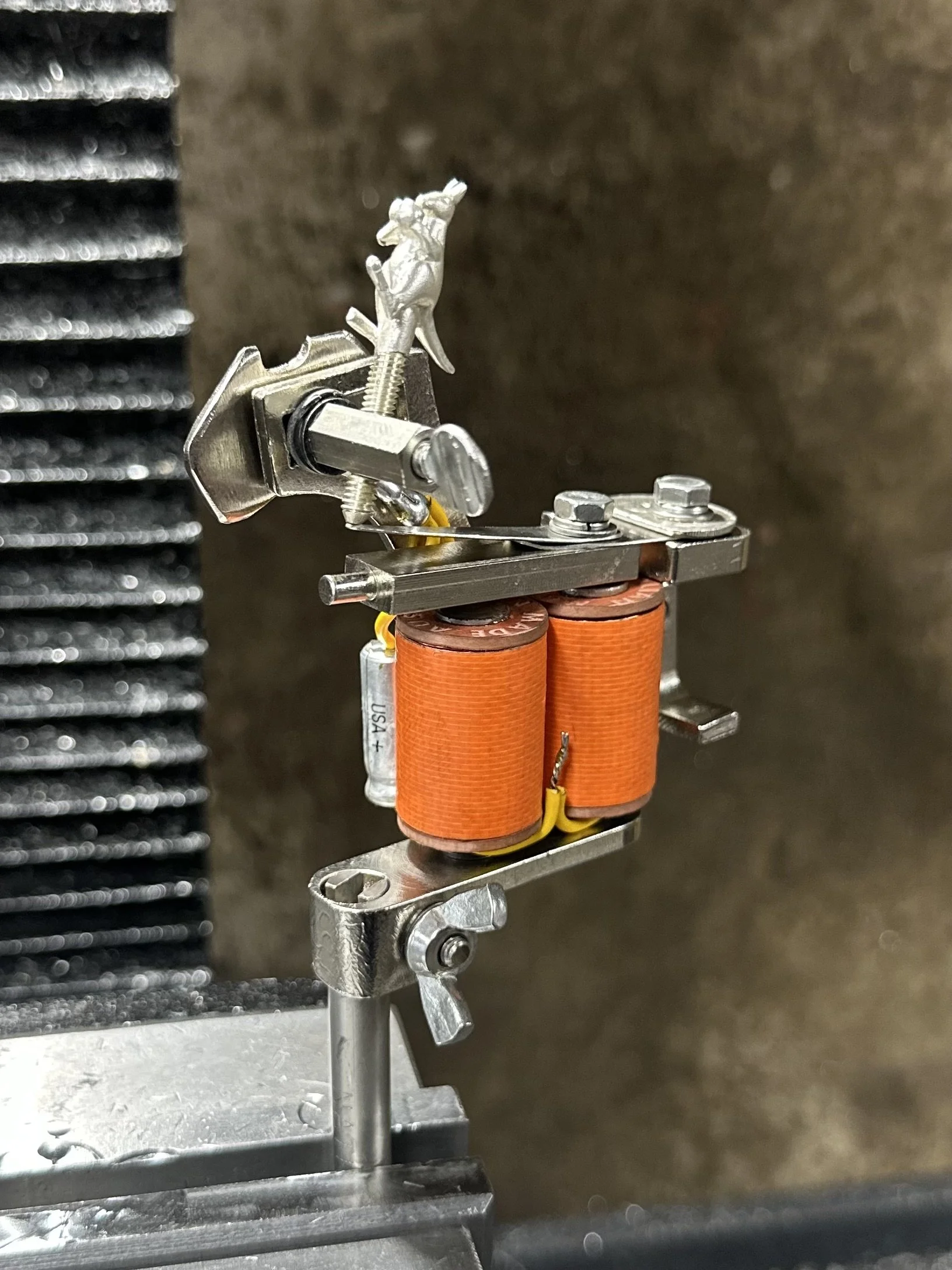 Close-up of a mechanical assembly with orange coils, metal parts, and a small figurine of a gray dinosaur on top, mounted on a metal base against a dark background.