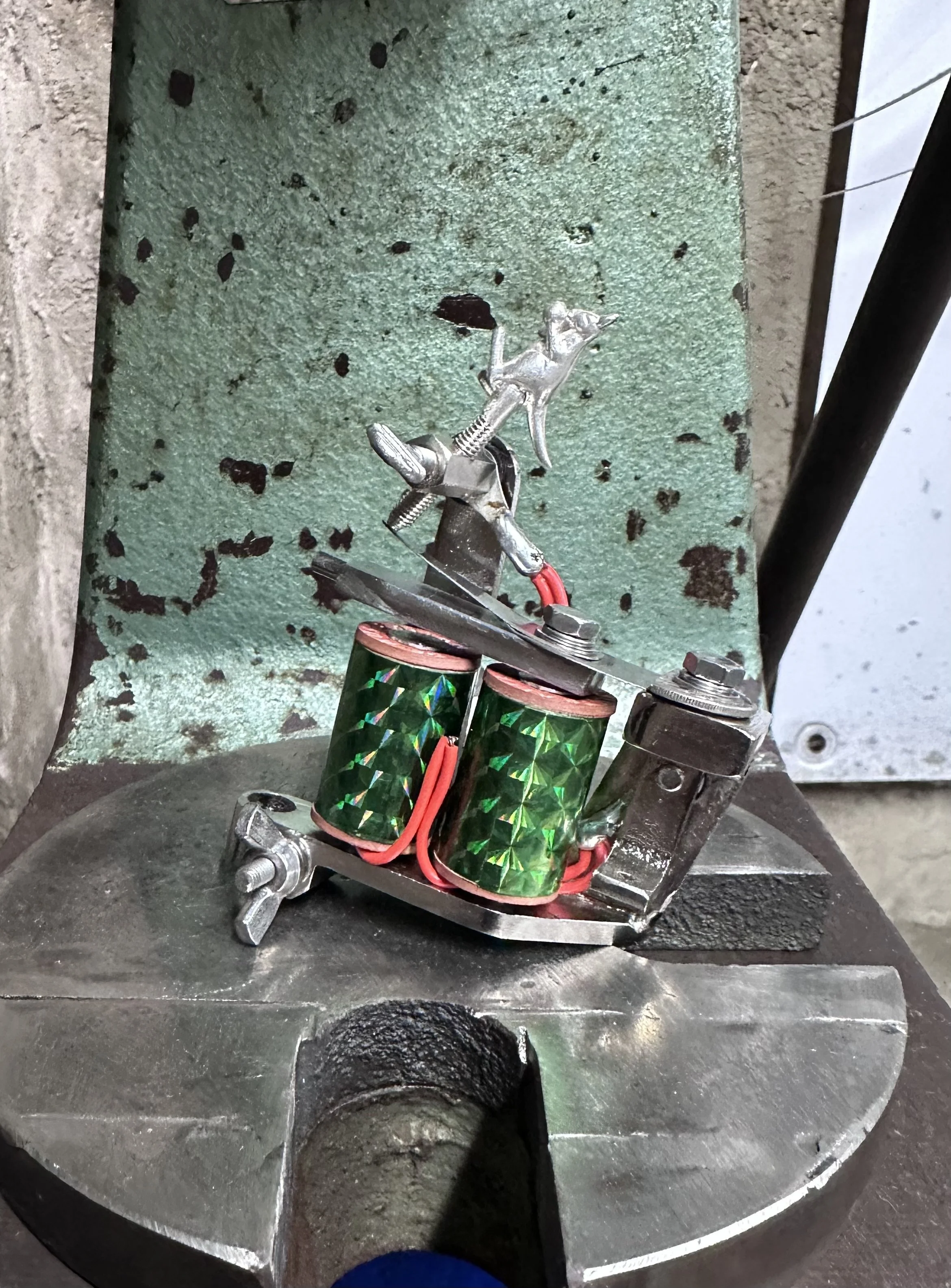 Close-up of a metallic coil with green and red wrapping, held by a metal clamp on a metallic surface, with a textured green and gray background.
