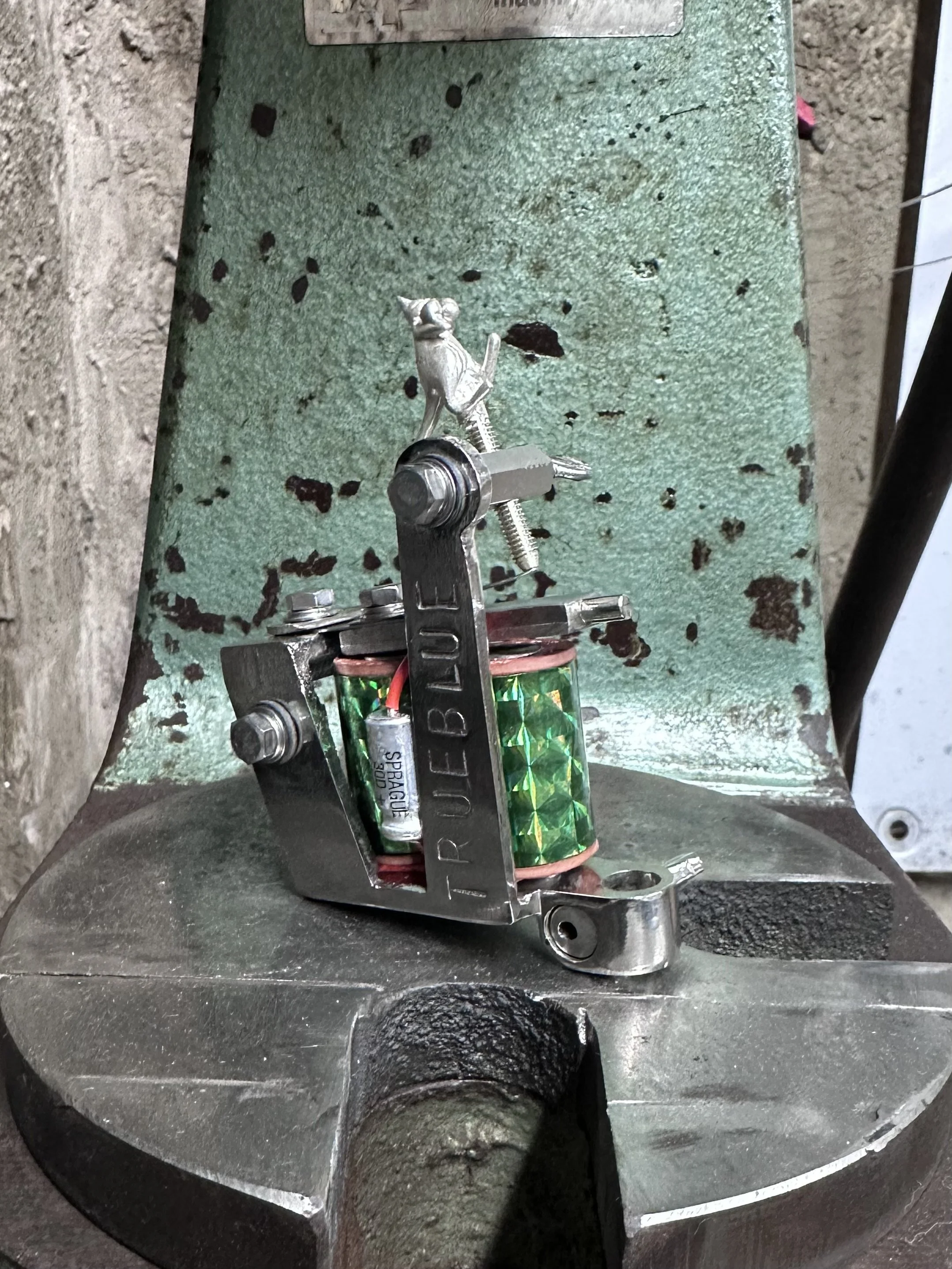 Close-up photo of a circuit setup with black wires, metal components, and a lithium battery, all mounted on a green textured surface, possibly part of a machine or device.