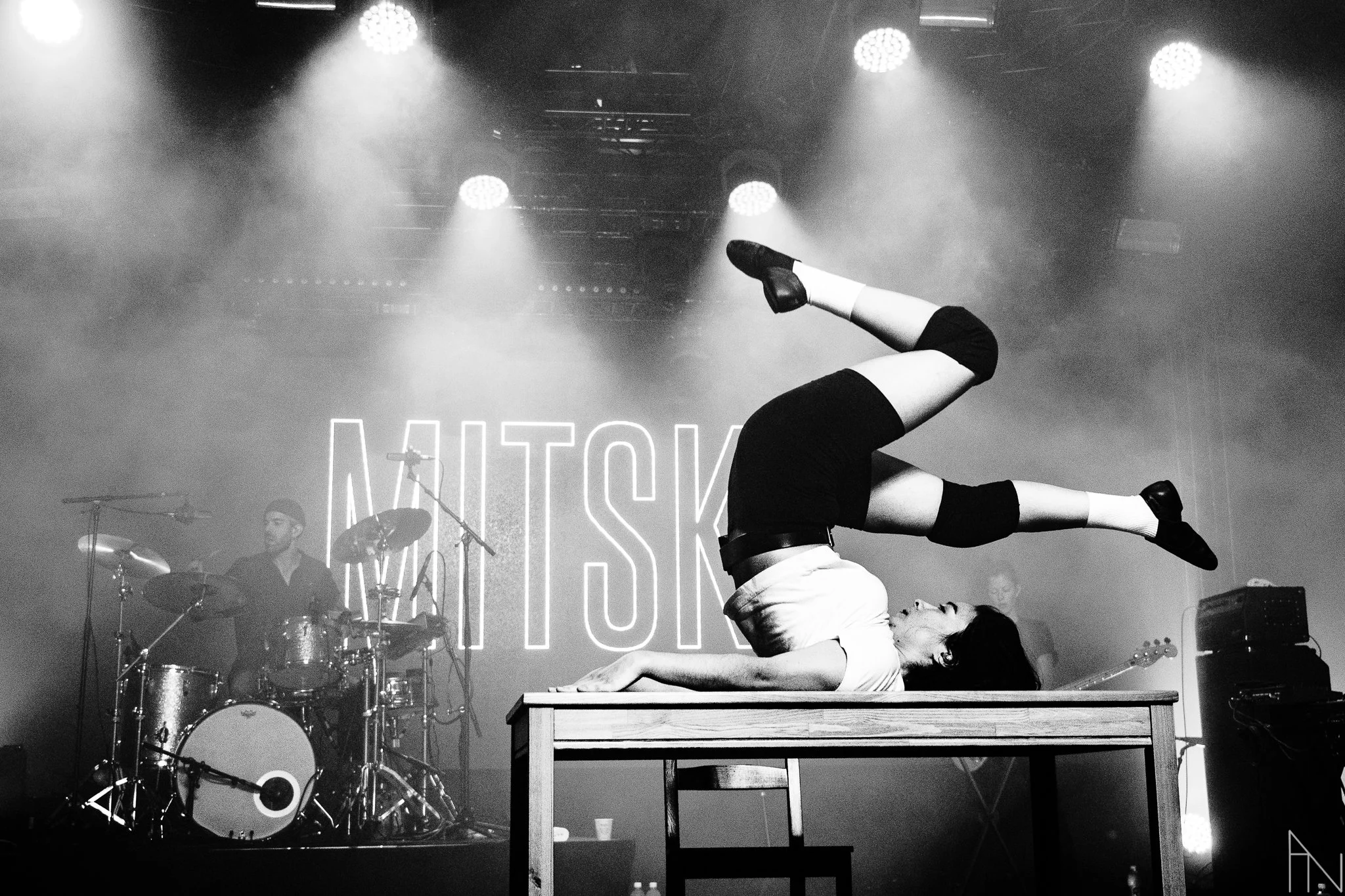 Mustavalkovalokuva henkilöstä, joka makaa pöydällä ja tekee voltin. Taustalla esiintyy bändi ja valoefektit, sekä teksti "MITSKI".