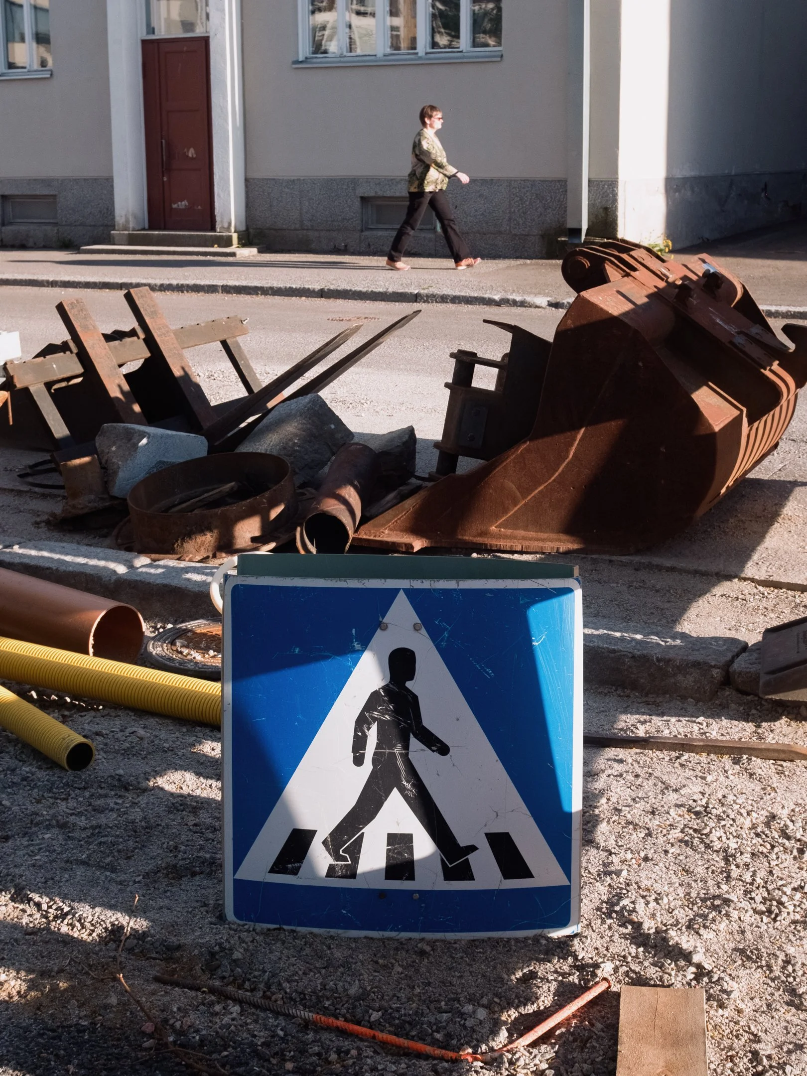 Kauppakatu tai tie, jossa on rakennustyömaa. Edessä on liikennemerkki, jossa on kävelevä ihminen ja suojatie. Taustalla on rakennustarvikkeita ja rautaa sekä kaupungin rakennusrappuinen seinä ja ikkunat.