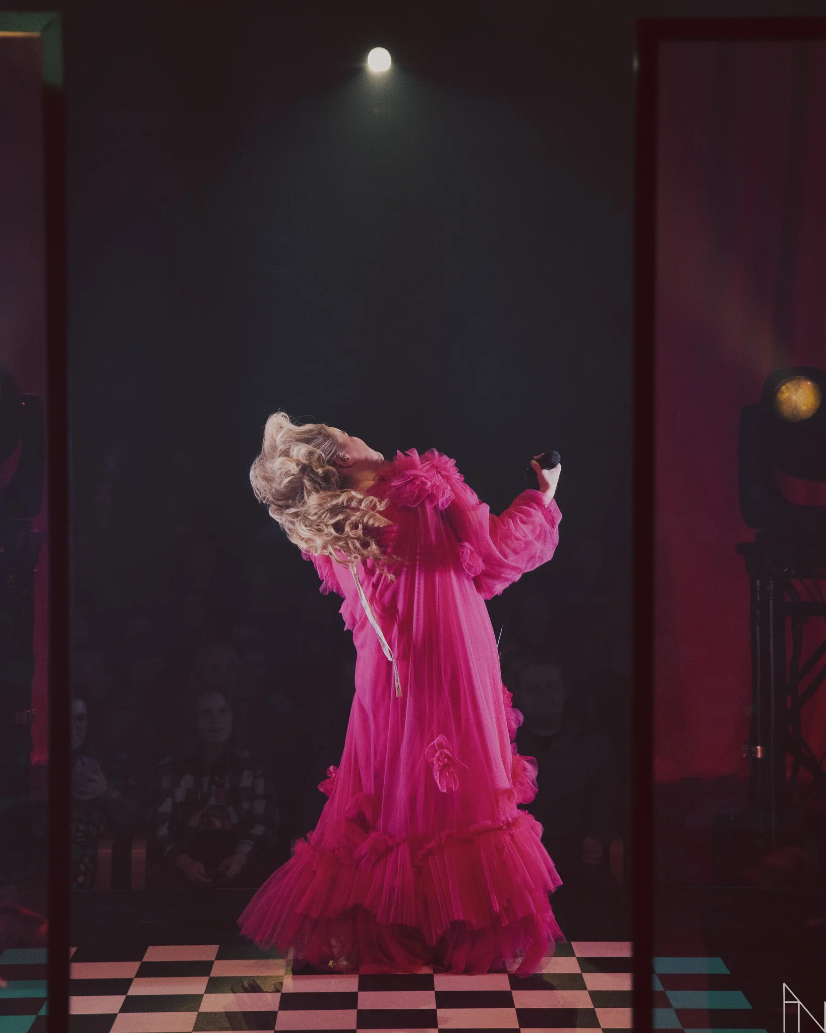 Naishahmo esiintyy lavalla kirkkaan pinkkinessä mekossa, joka on koristeltu kankailla ja röyhelöillä. Hän pitää mikrofonia kädessään ja ehkä laulaa tai puhuu. Taustalla on yleisö ja lavavalot.