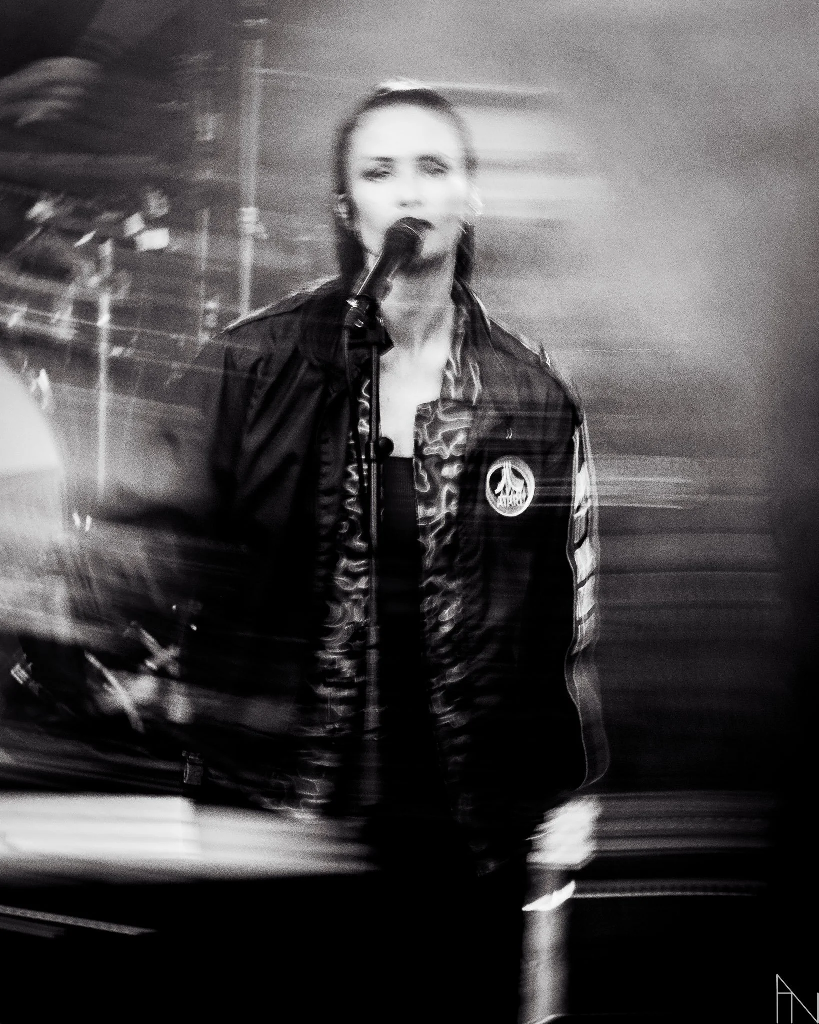 Mustavalkoinen kuva naisesta, joka puhuu mikrofoniin ja on pukeutunut pariin vaatteeseen, yksi niistä on iloisen pehmeä markiisikuvioinen jakku. Taustalla on sumeita ja liikevaikutteisia valoja ja rakenteita.