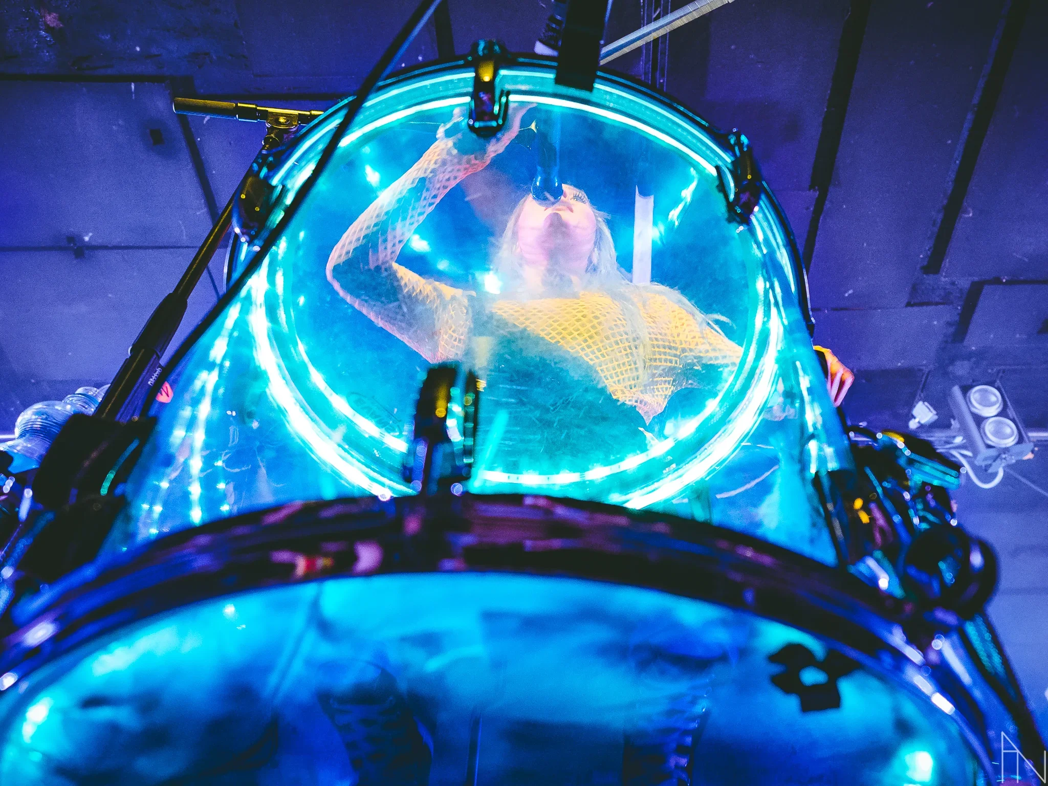 Naisartisti kuvattu läpinäkyvän rummun läpi, jota ympäröi siniset ja neonvalot, suurikokoinen LED-valo-ympyrä ympärillä.