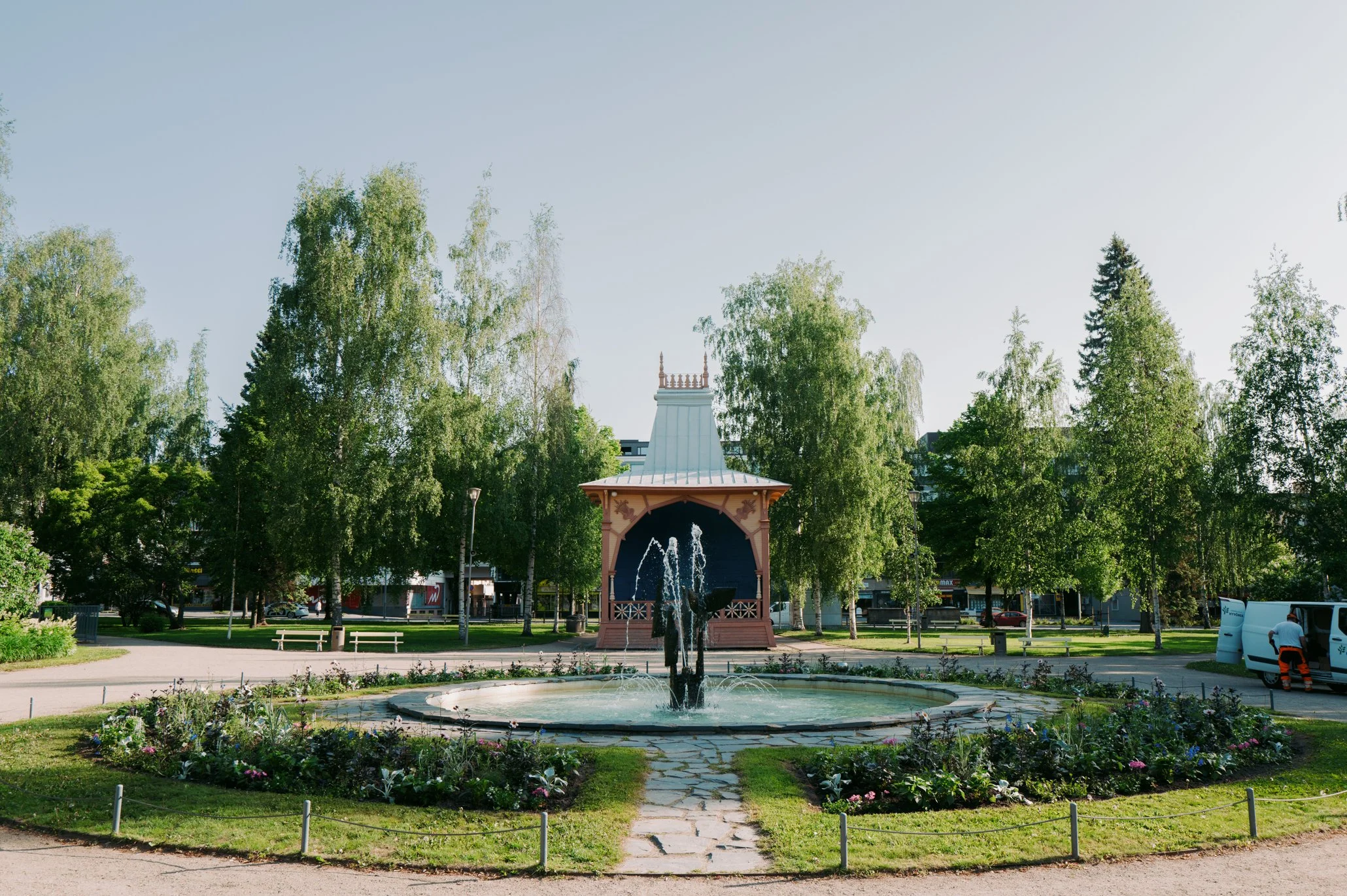 Kukkaistutuksia ja vesilähde kaupunginpuistossa, taustalla double decker rakennus ja saaria muistuttava torni, vihreitä puita ja selkeä taivas. Mikkelin kirkkopuisto.