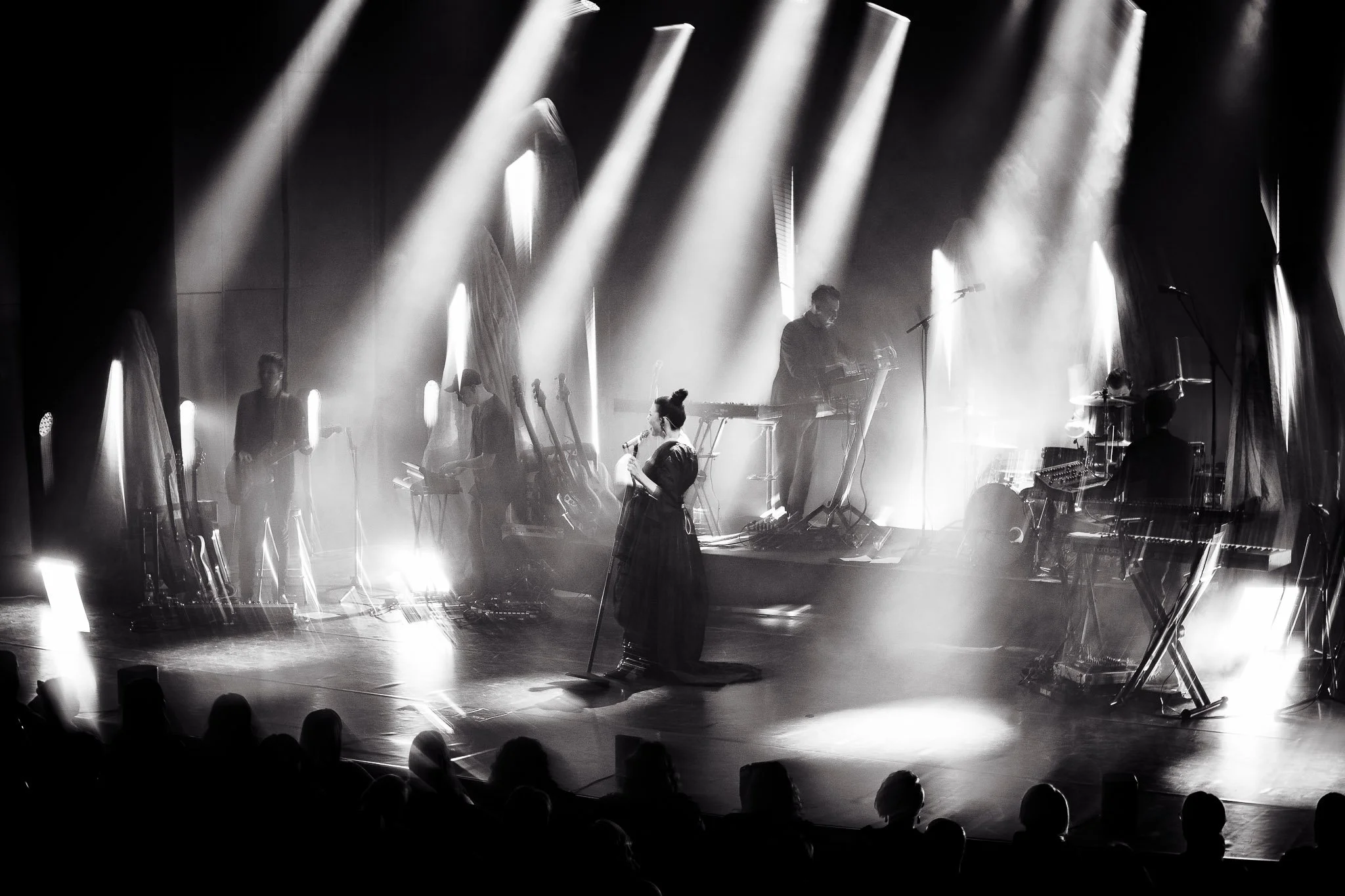 Mustavalkoinen kuva musiikkiesityksestä, jossa useita soittajia lavalla ja lavasteisiin suunnatut valot, yleisön siluetteja etualalla.