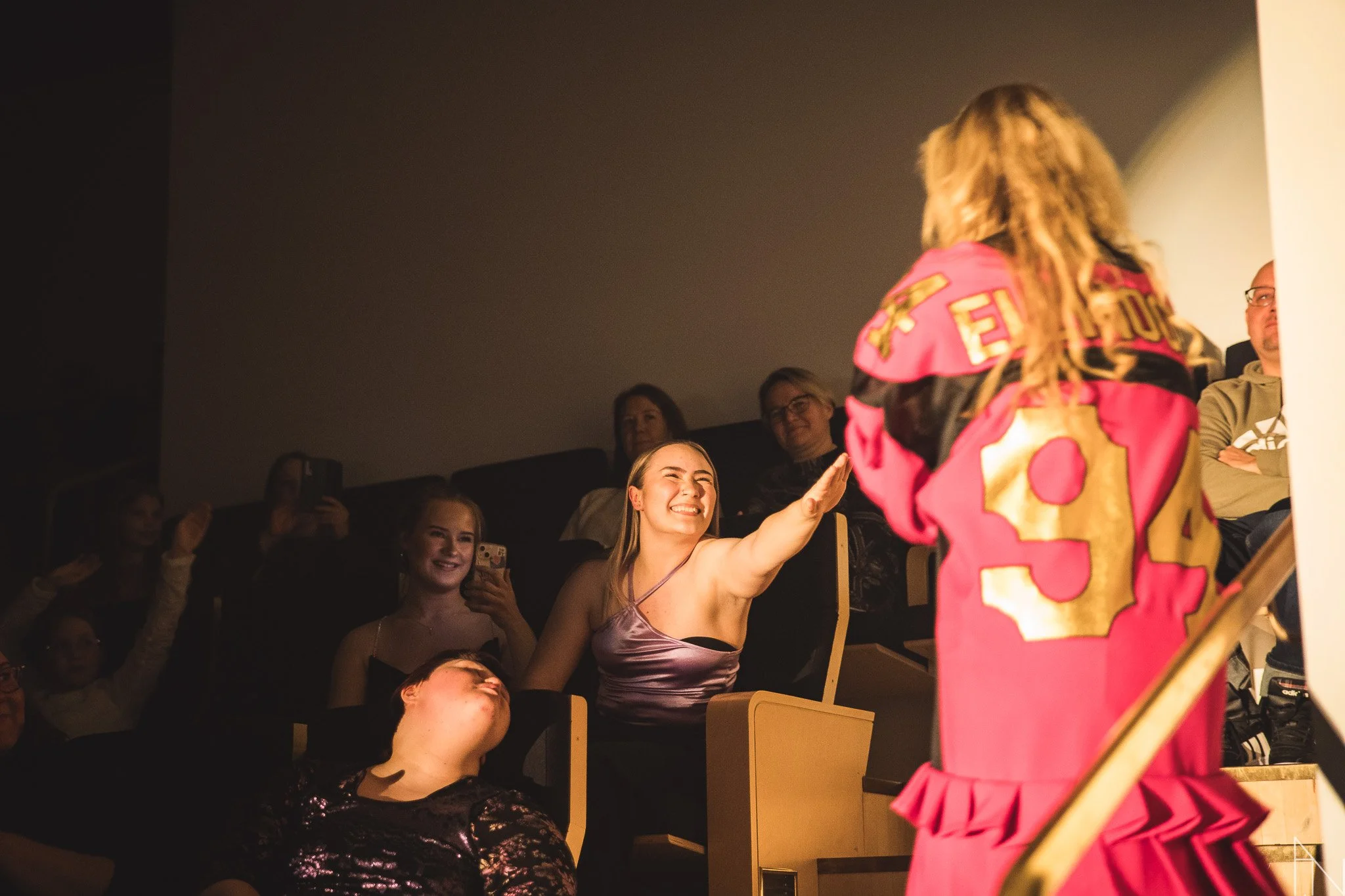 Nainen hymyilee ja ojentaa kättään yleisön edessä, jossa ihmiset ottavat kuvia ja katsovat kiinnostuneina. Ellinoora Mikaeli 2024.