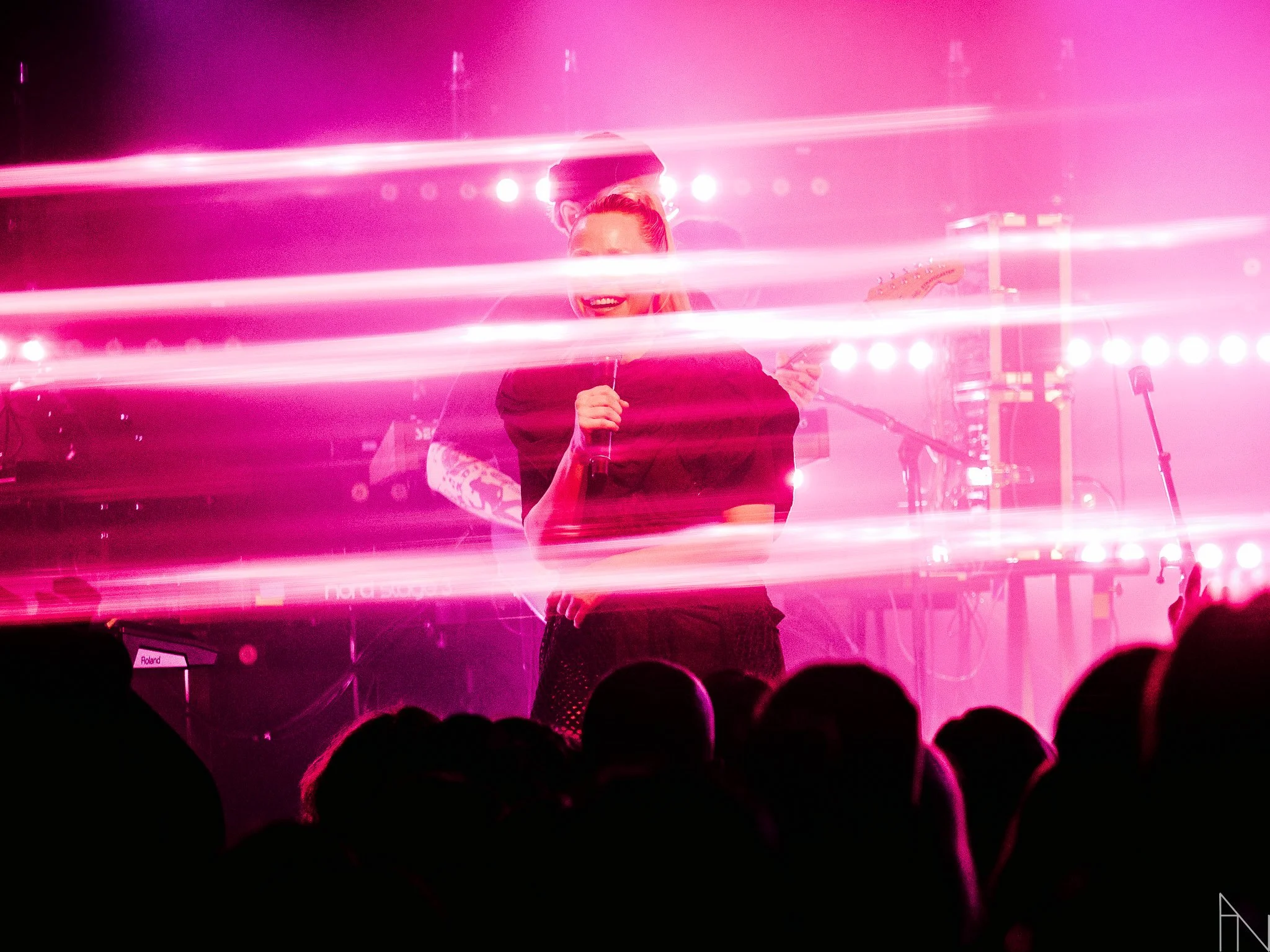 Nainen laulamassa lavalla, taustalla instrumentteja, edessä valoefektejä ja yleisöä, pinkit ja violetit valaistus.