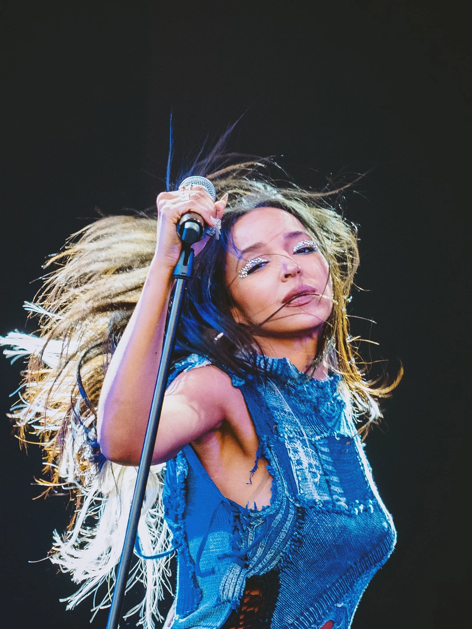 Nainen laulamassa mikrofonilla, hulmuavat hiukset, musta tausta, vaatteet sininen ja farkkuinen, näyttävästi meikattu. Tinashe.