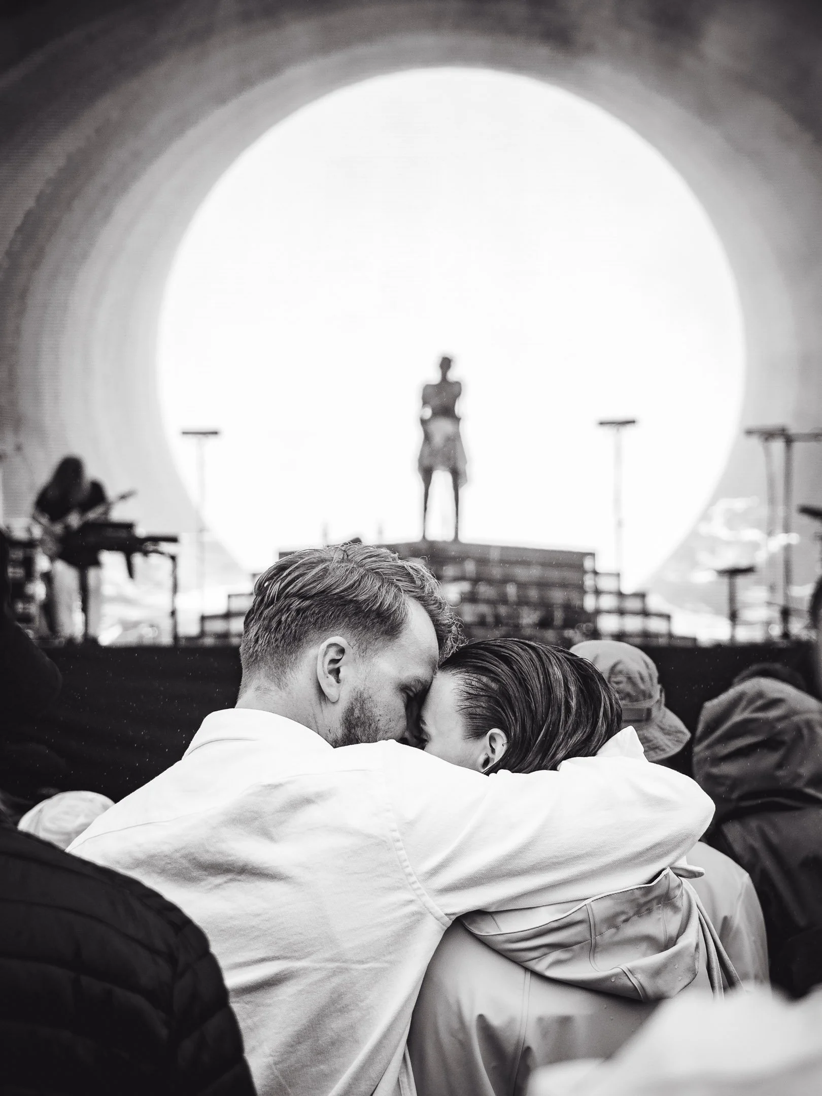 Kaksi ihmistä halaavat siten, että heidän otsansa ovat kosketuksissa, ja heidän silmänsä ovat suljettuina. Taustalla näkyy näyttävä suuri valkoinen pyöreä rakenne ja konserttilava.