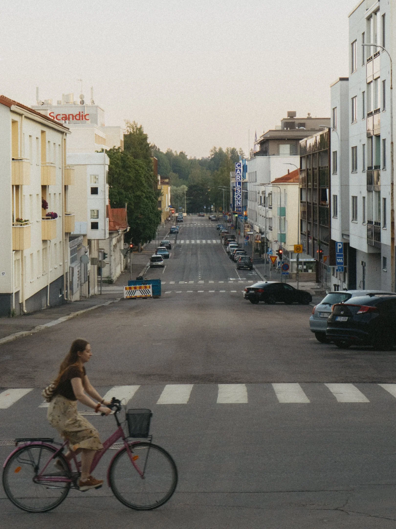 Kuva Mikonkadulta Mikkelissä, jossa on rivissä autoja ja rakennuksia. Etualalla nainen polkee pyörällä; taustalla on puita ja rakennuksia, kuten hotelli Scandic ja Carlsson-kauppakatu.